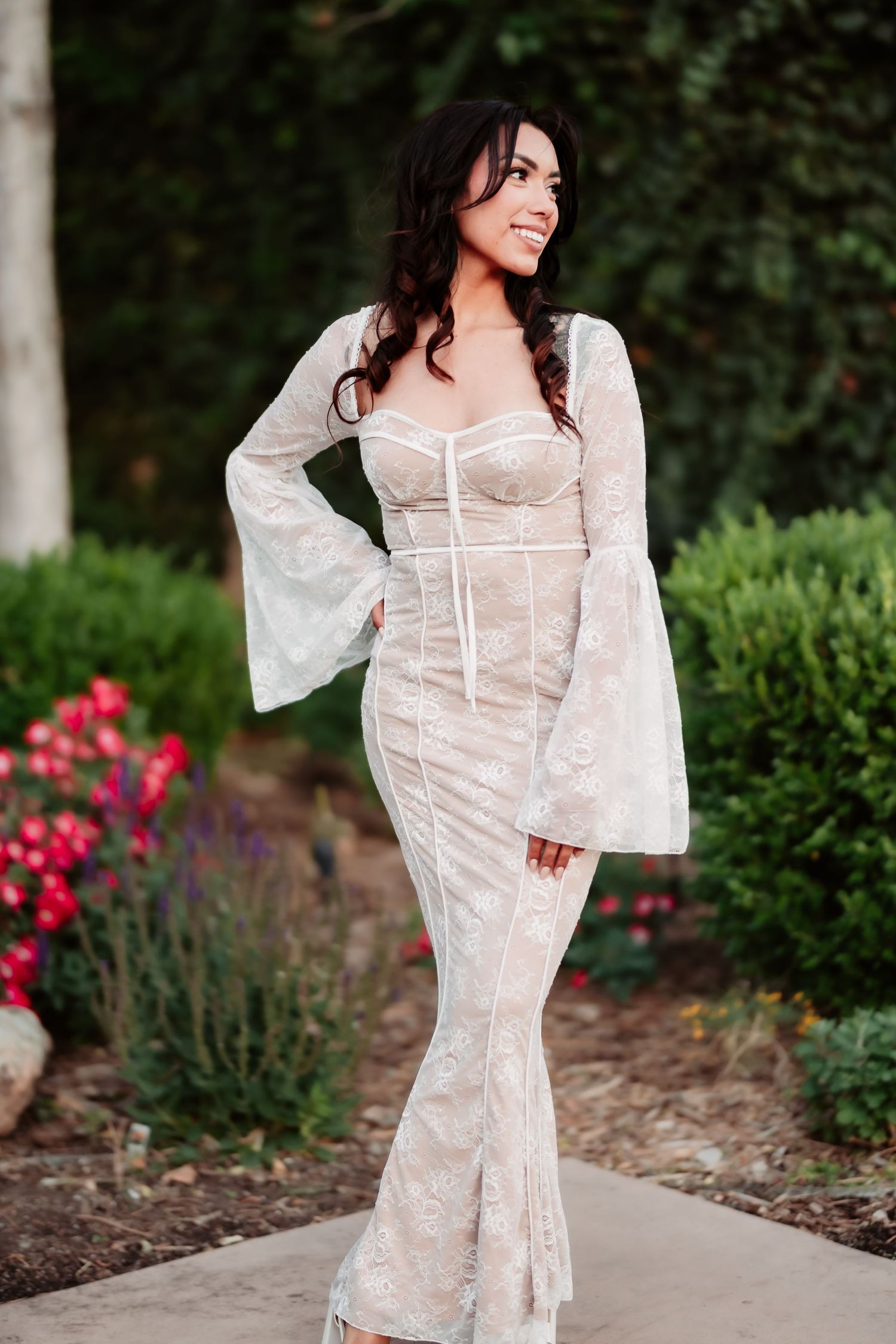 Woman poses in white lace dress against garden backdrop with blooming flowers.
