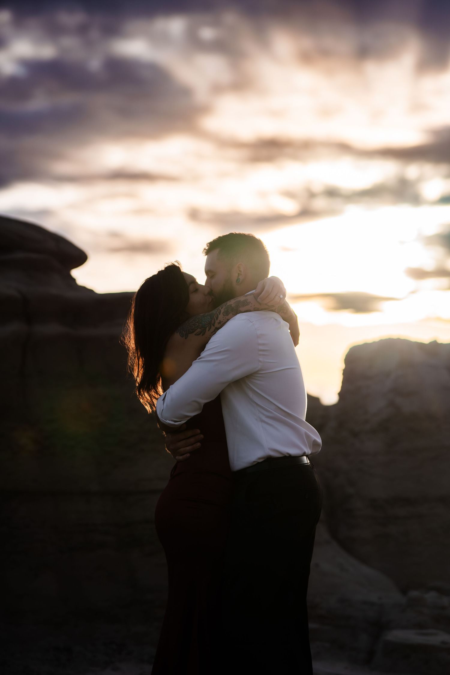 Silhouetted couple shares intimate moment against dramatic sunset sky in rocky desert terrain.
