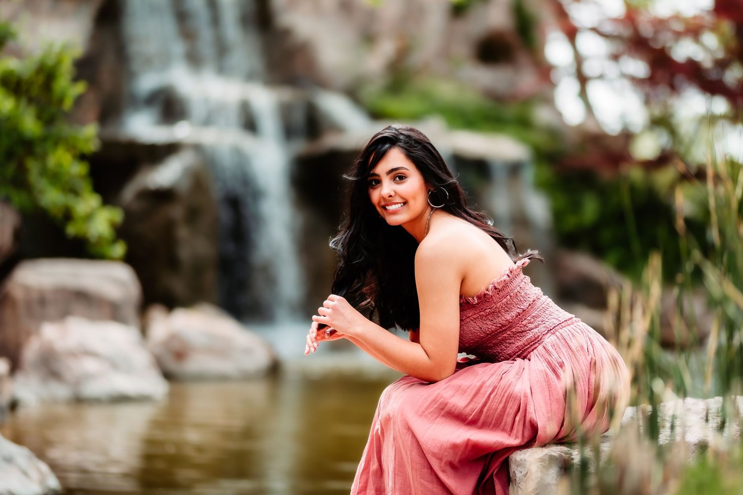 A woman in a flowing pink dress poses gracefully by a waterfall in a lush garden setting.