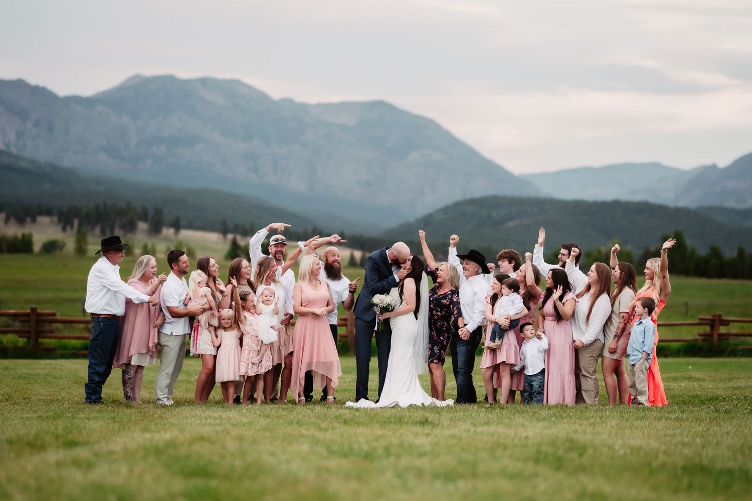 Large wedding party poses in coordinated attire against mountain meadow backdrop.