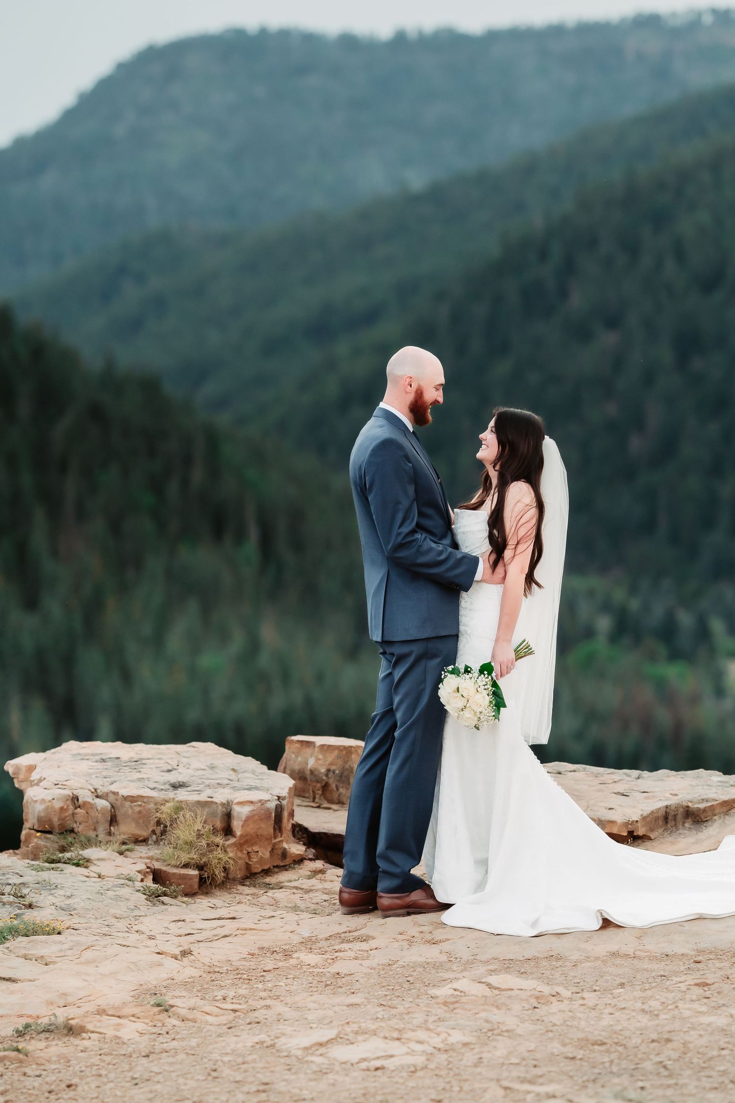 Newlyweds share tender moments on rocky mountain overlook with expansive forest views.