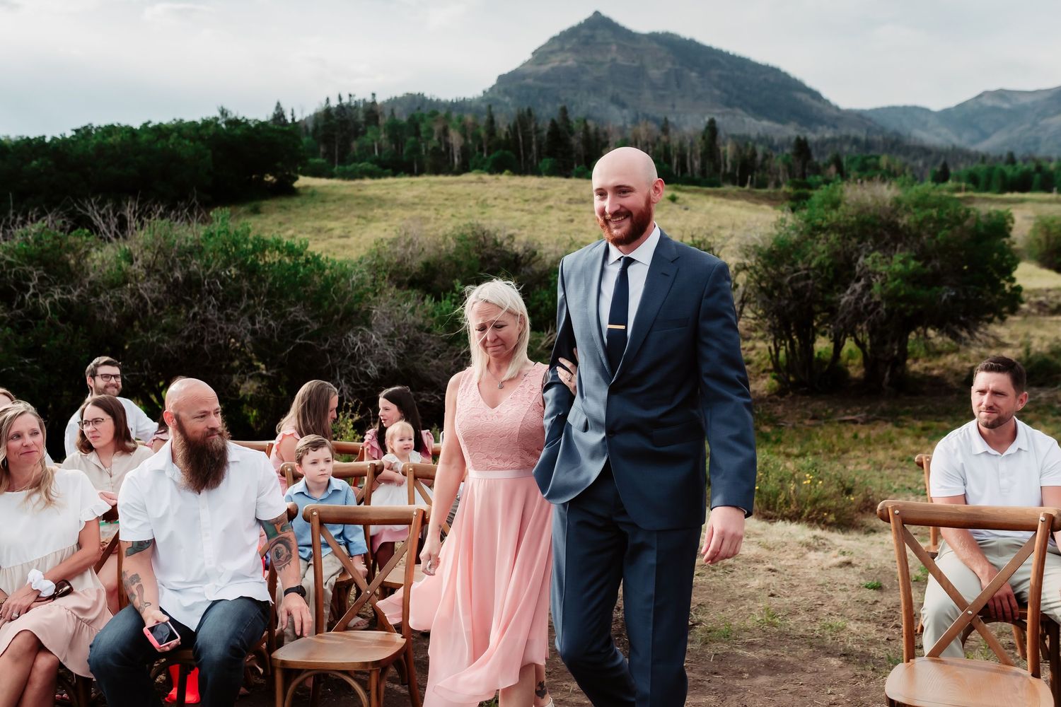 Outdoor mountain wedding ceremony with guests seated on wooden chairs in a scenic natural setting.