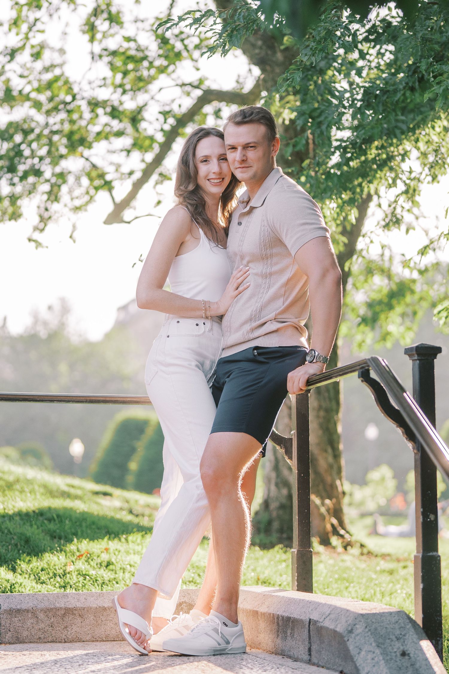 Two people posing together on stone steps in a park setting during golden hour, surrounded by green trees.