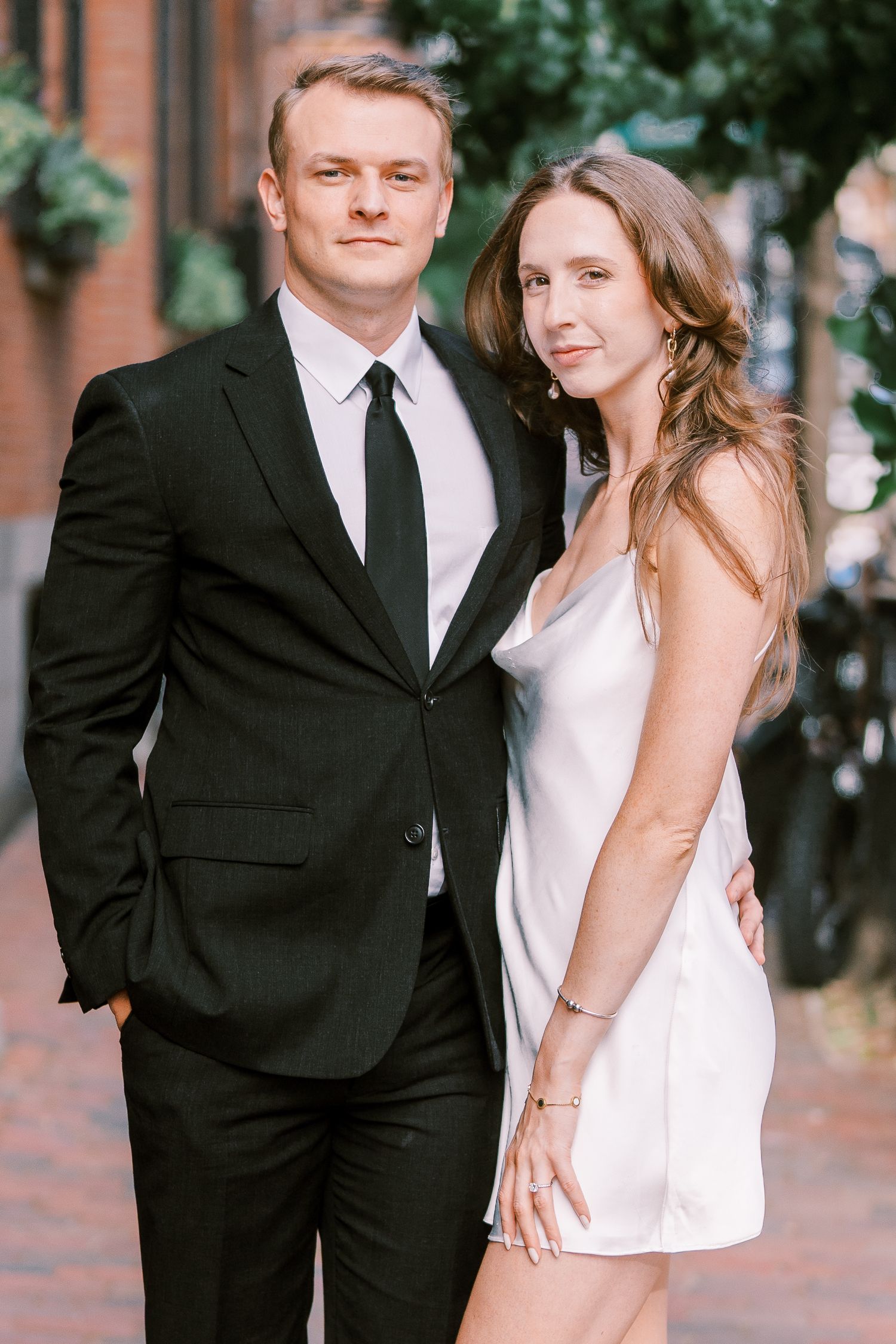 Couple in formal attire poses together on brick sidewalk for romantic engagement or wedding photos.