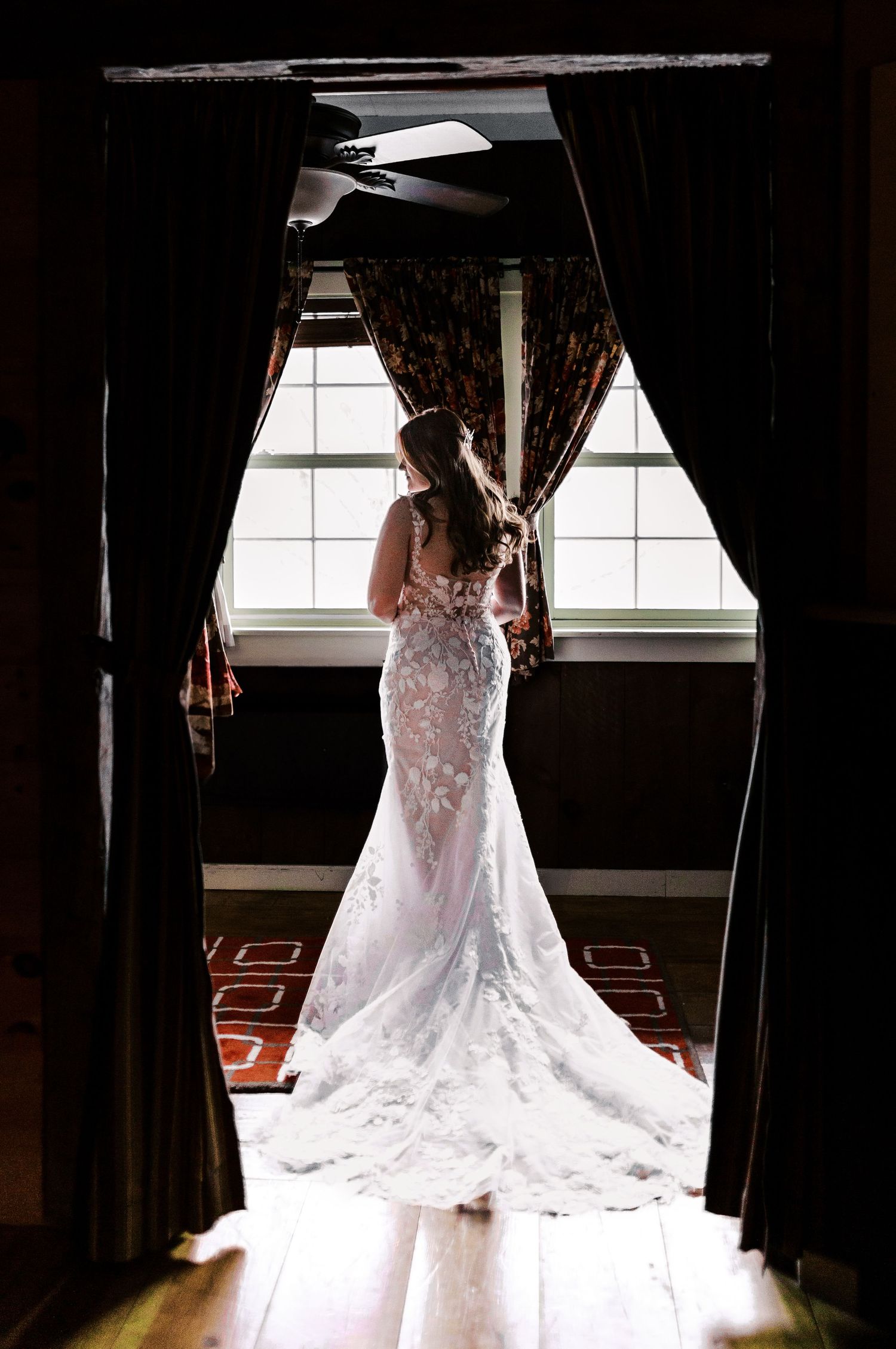 Silhouette of a bride in a fitted lace wedding gown standing by a window with flowing curtains.
