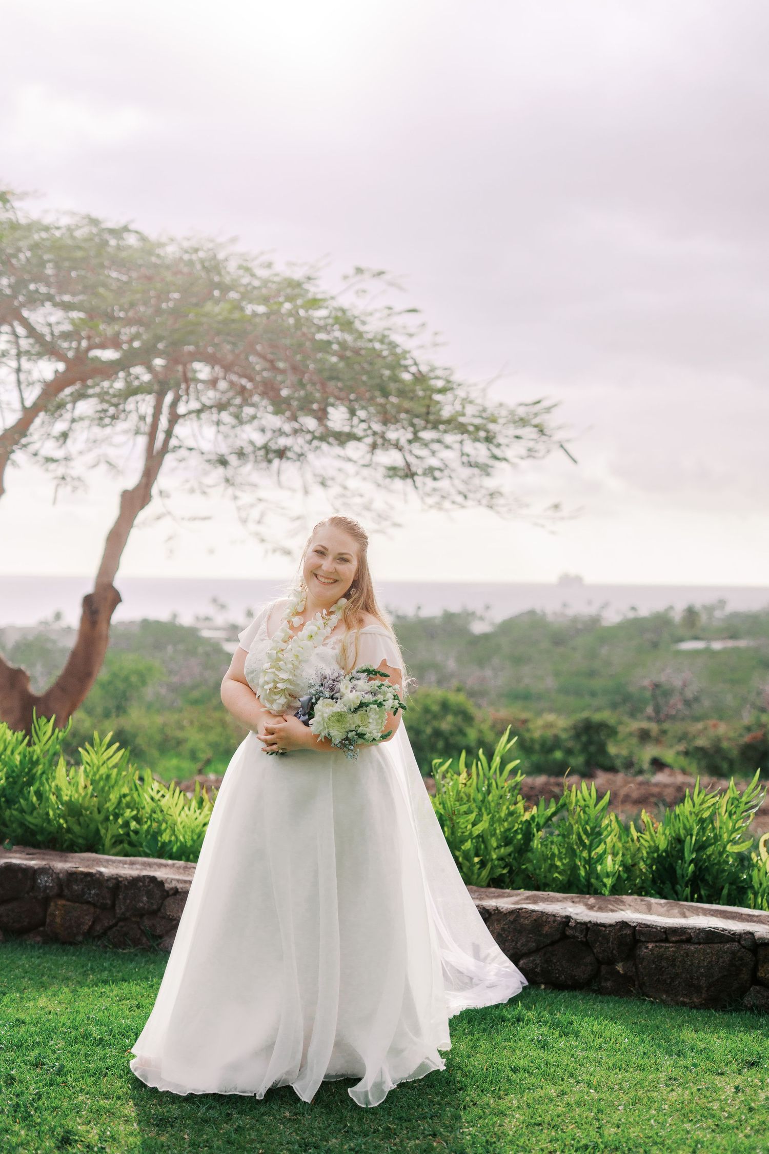 Wedding photography of a person in a flowing white gown holding a bouquet in a scenic outdoor garden setting.