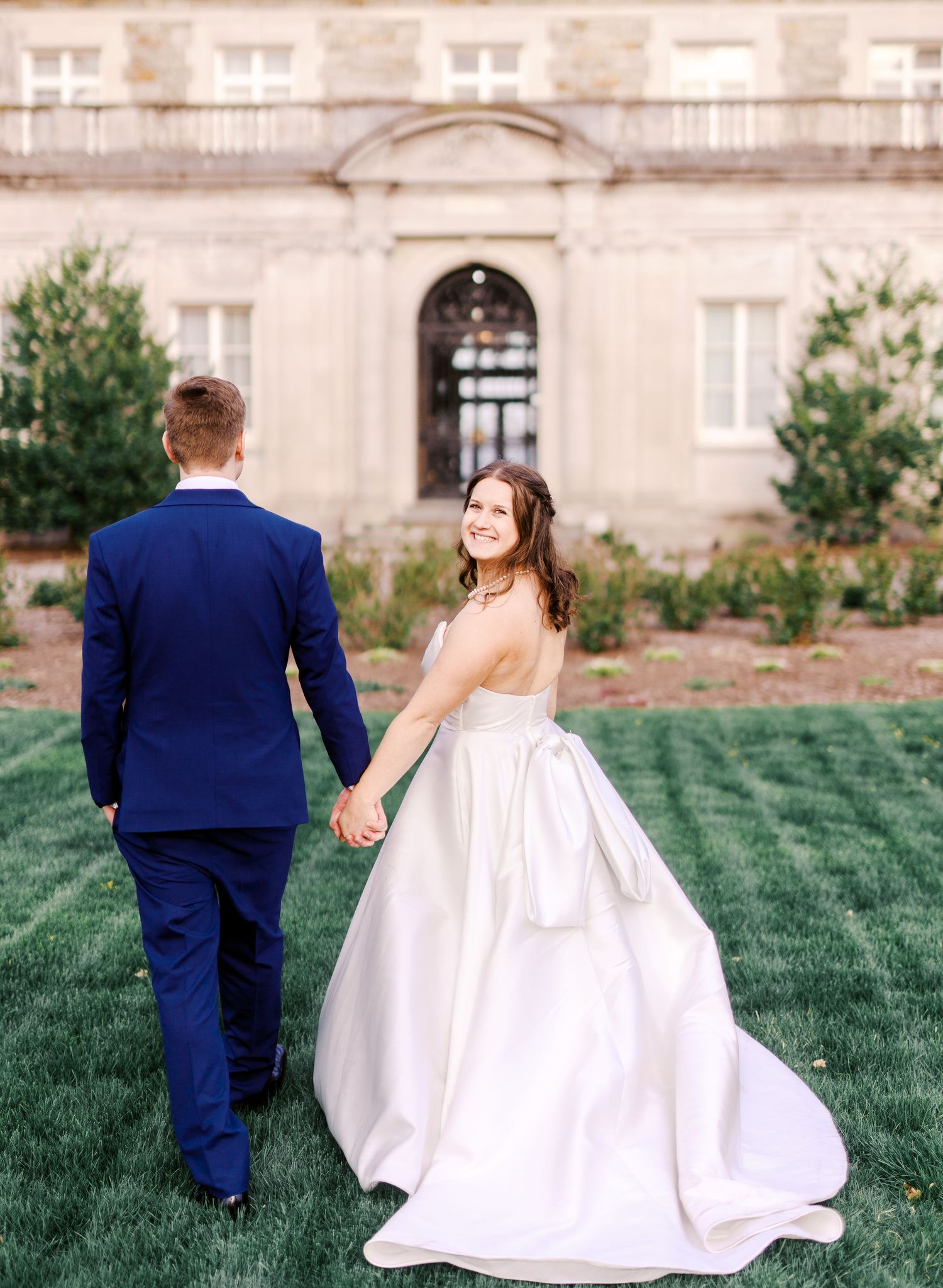 A bride and groom walking on manicured lawn towards elegant stone mansion entrance.