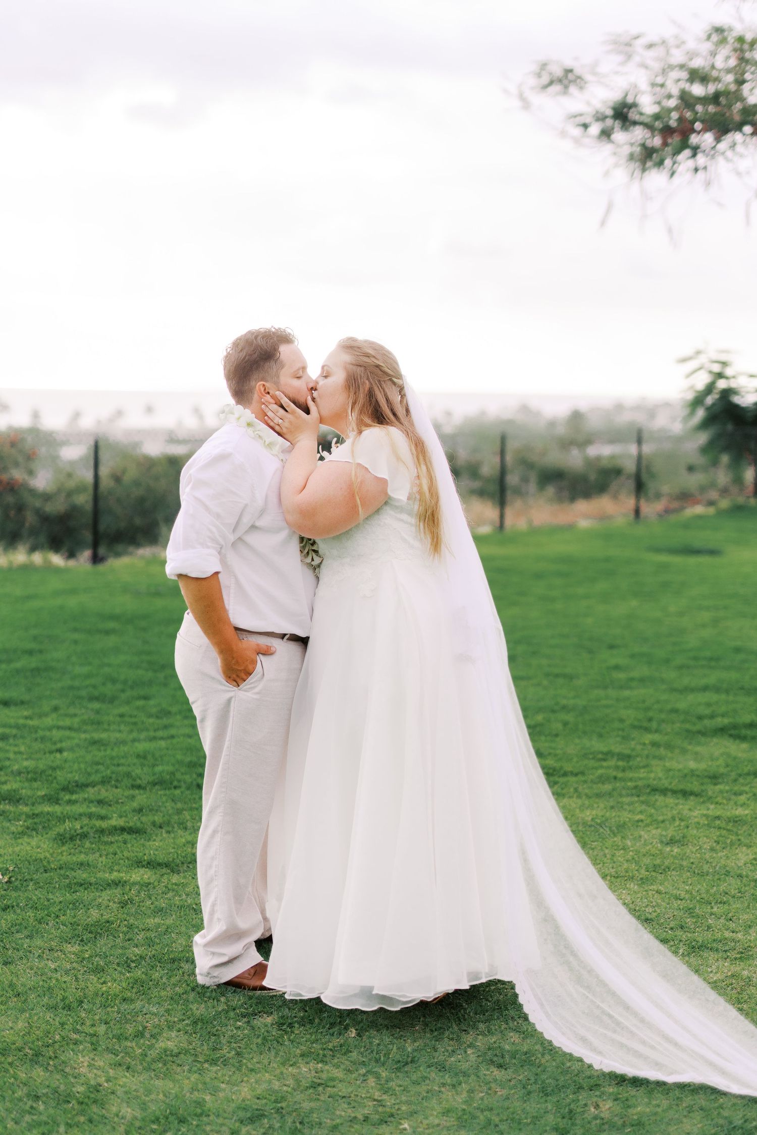 A couple shares their first kiss as newlyweds on a grassy hill overlooking a city.