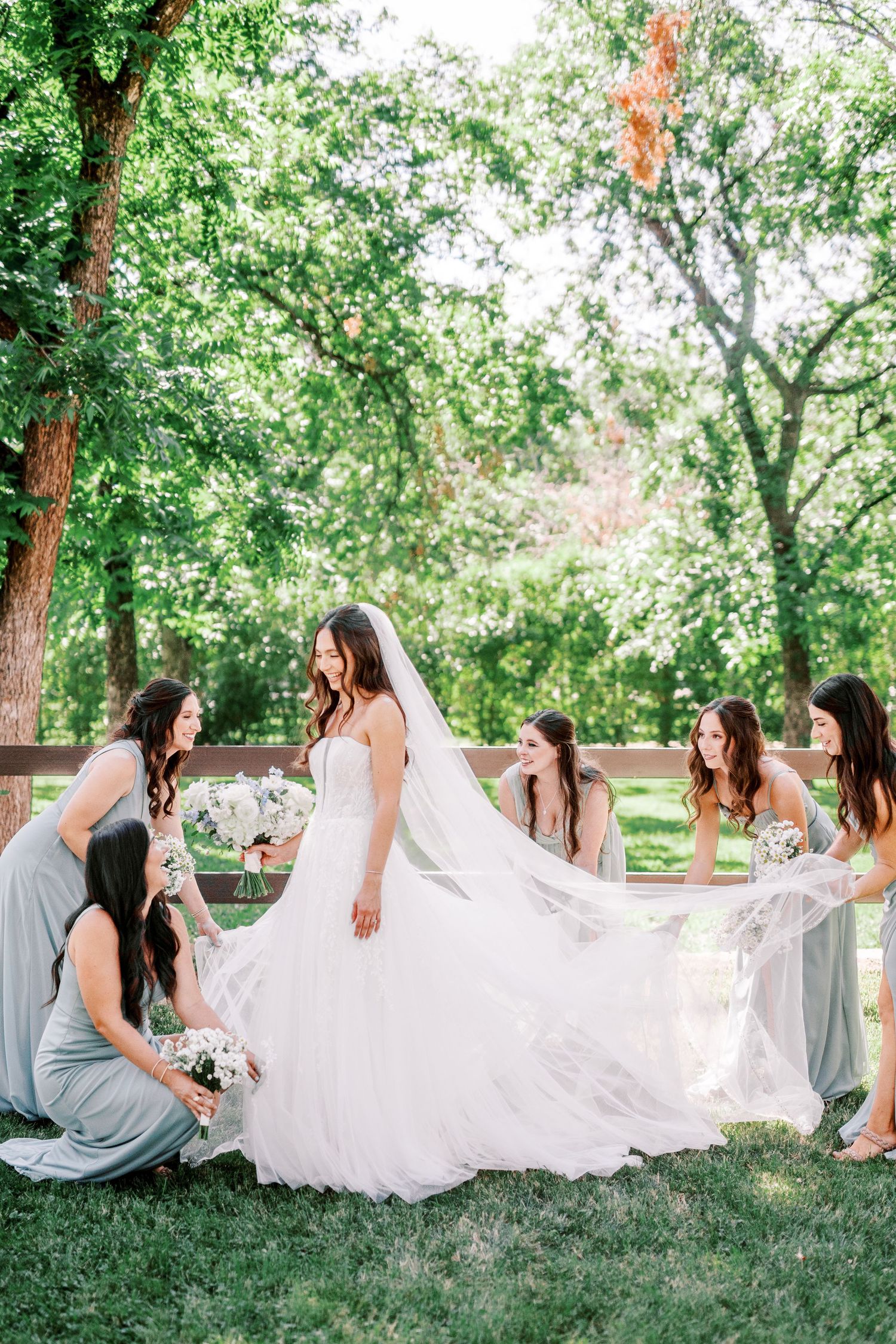 Group wearing grey bridesmaid dresses gather around bride in flowing white gown and veil in outdoor garden setting.