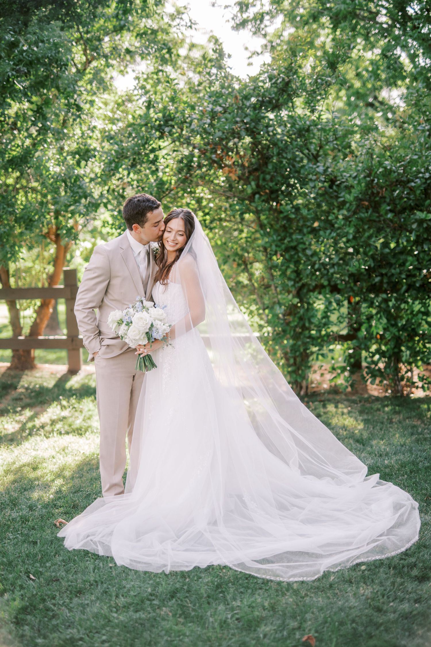 A romantic outdoor wedding scene with a couple in light beige and white attire standing on lush green grass beneath trees.