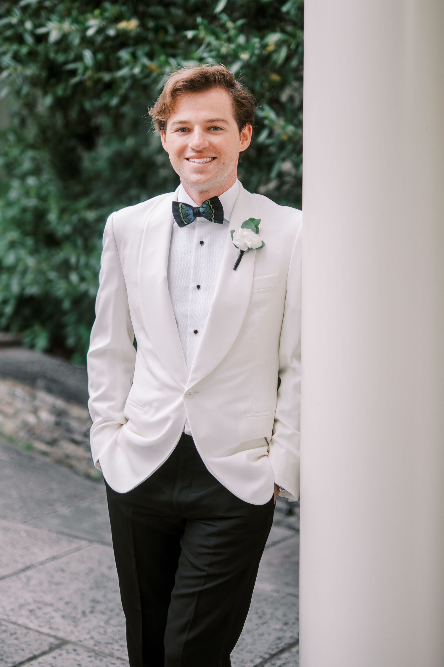 A groom in a classic white tuxedo jacket and black pants poses elegantly against white columns in an outdoor setting.