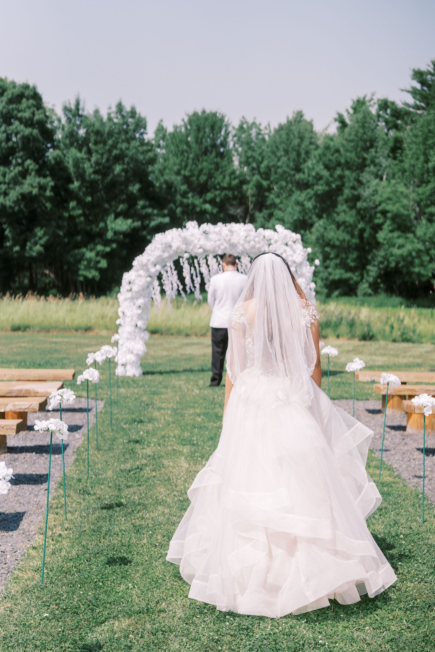 A white balloon arch frames an outdoor wedding ceremony setup with a bride in a flowing gown on a grassy lawn.