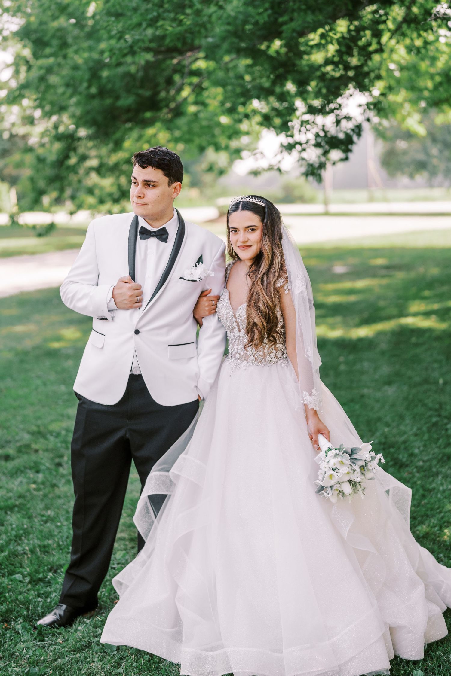 A bride and groom pose together outdoors in a park setting, with the bride wearing a flowing white gown.