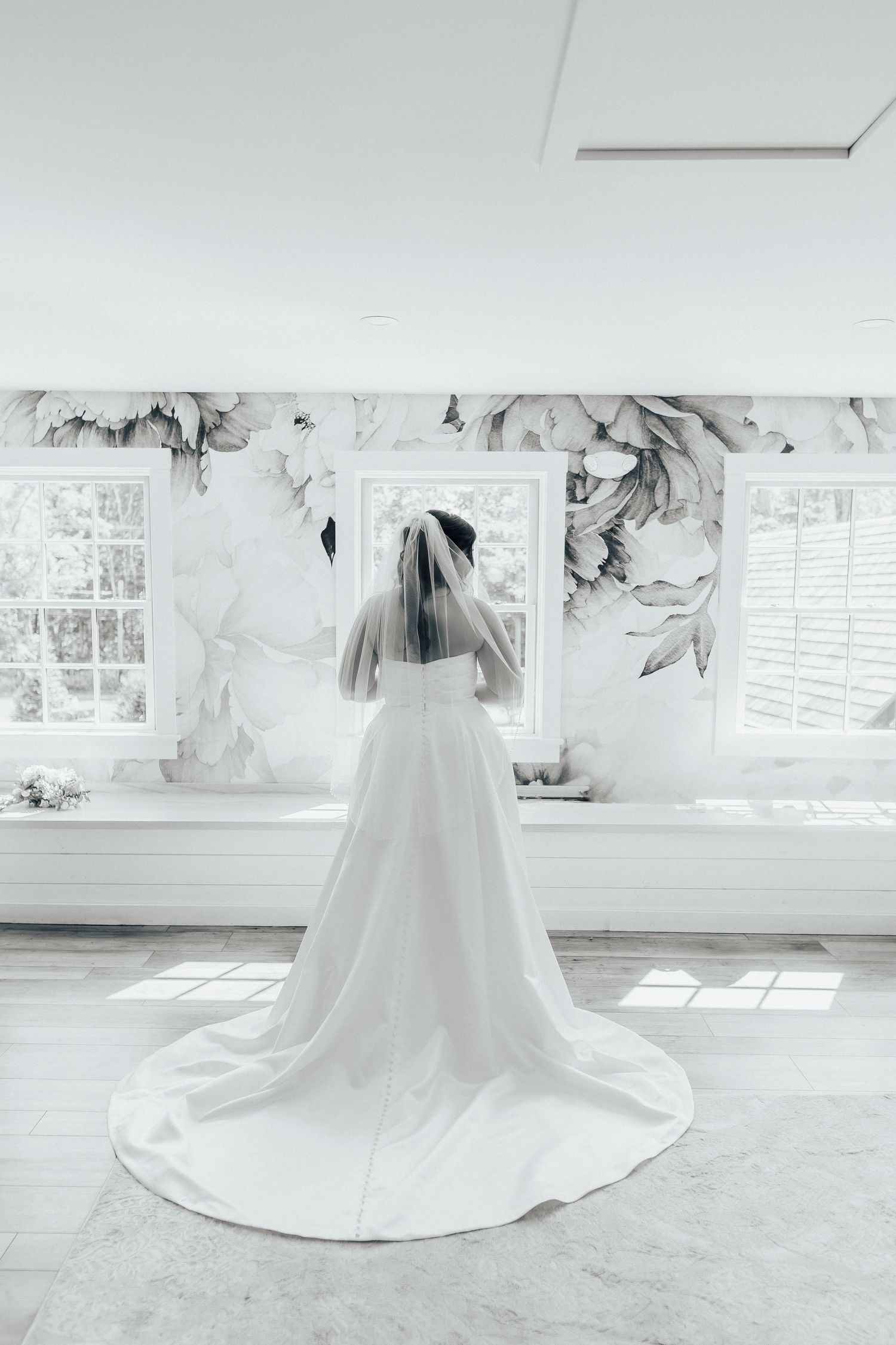 Wedding dress with long train photographed from behind in a bright white room with floral wallpaper.