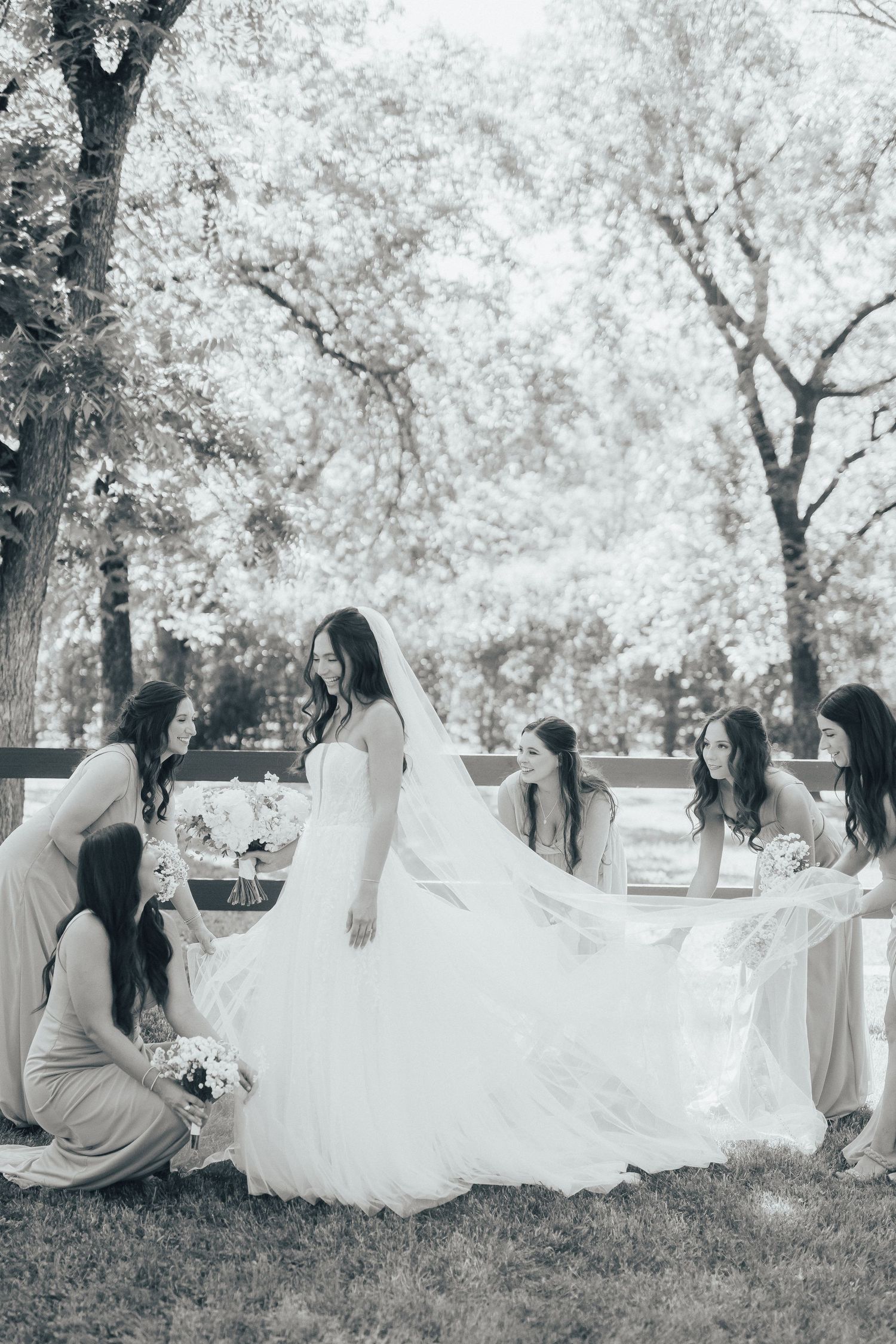 A wedding party gathers on a lawn with bridesmaids helping arrange the bride's flowing white dress train.