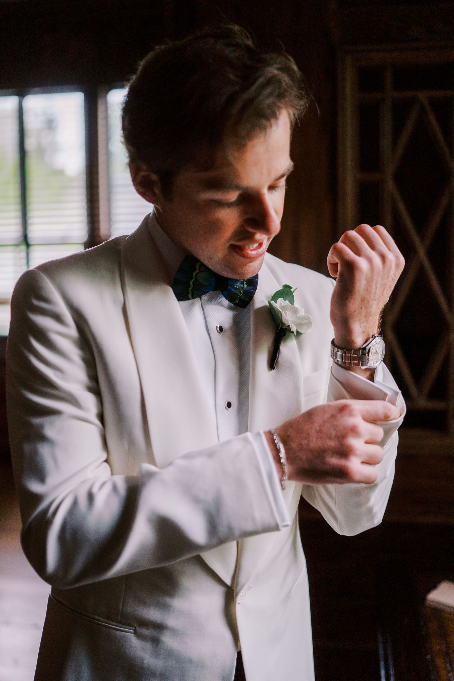 A groom in a white tuxedo and bowtie makes final adjustments to his cufflinks in the atmospheric room lighting.