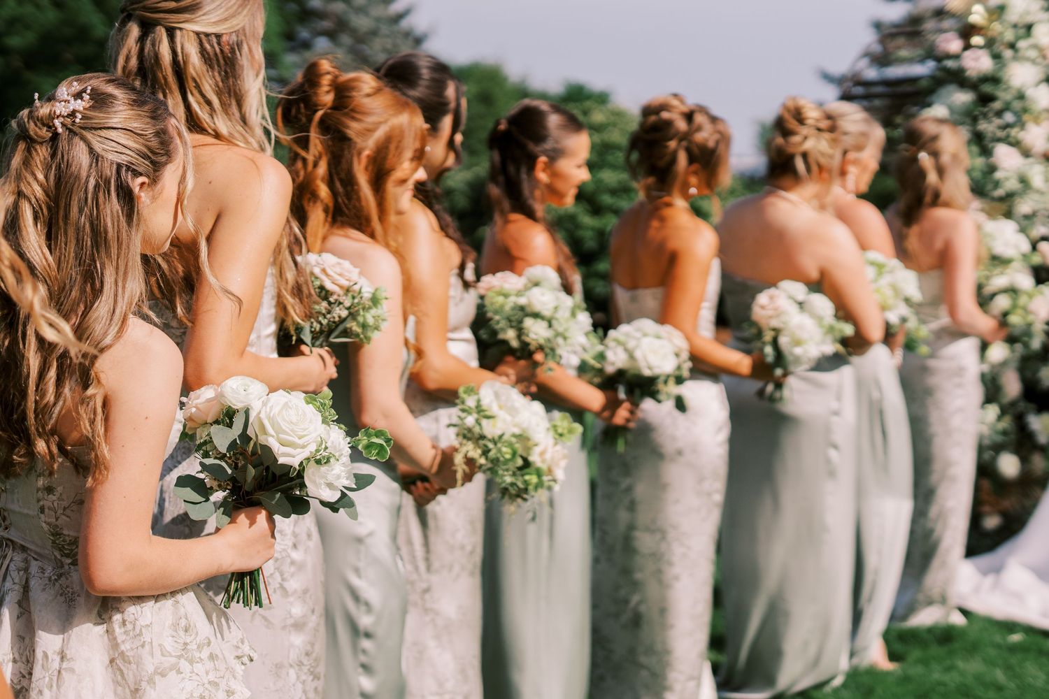 Bridesmaids in matching light-colored dresses hold white floral bouquets while standing in a row at an outdoor wedding.