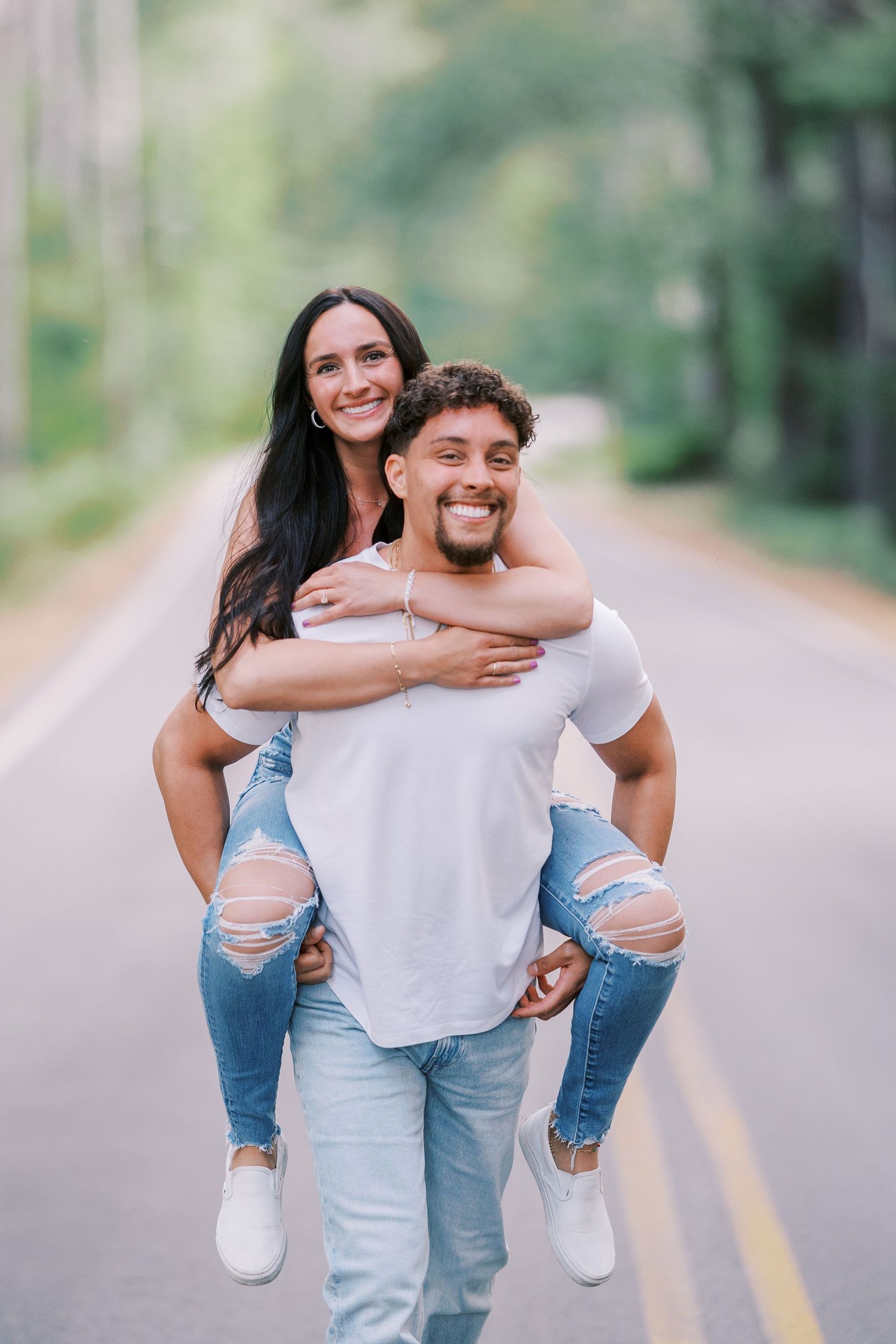 Joyful couple having fun with piggyback ride on an empty road surrounded by trees.
