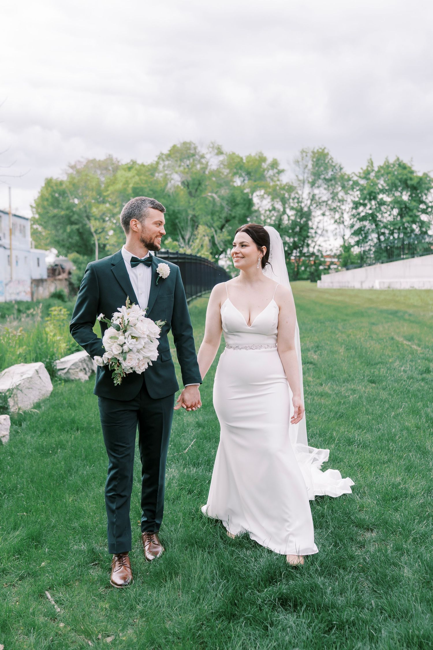 Newlyweds walking hand in hand on grassy field with bride in fitted white gown.