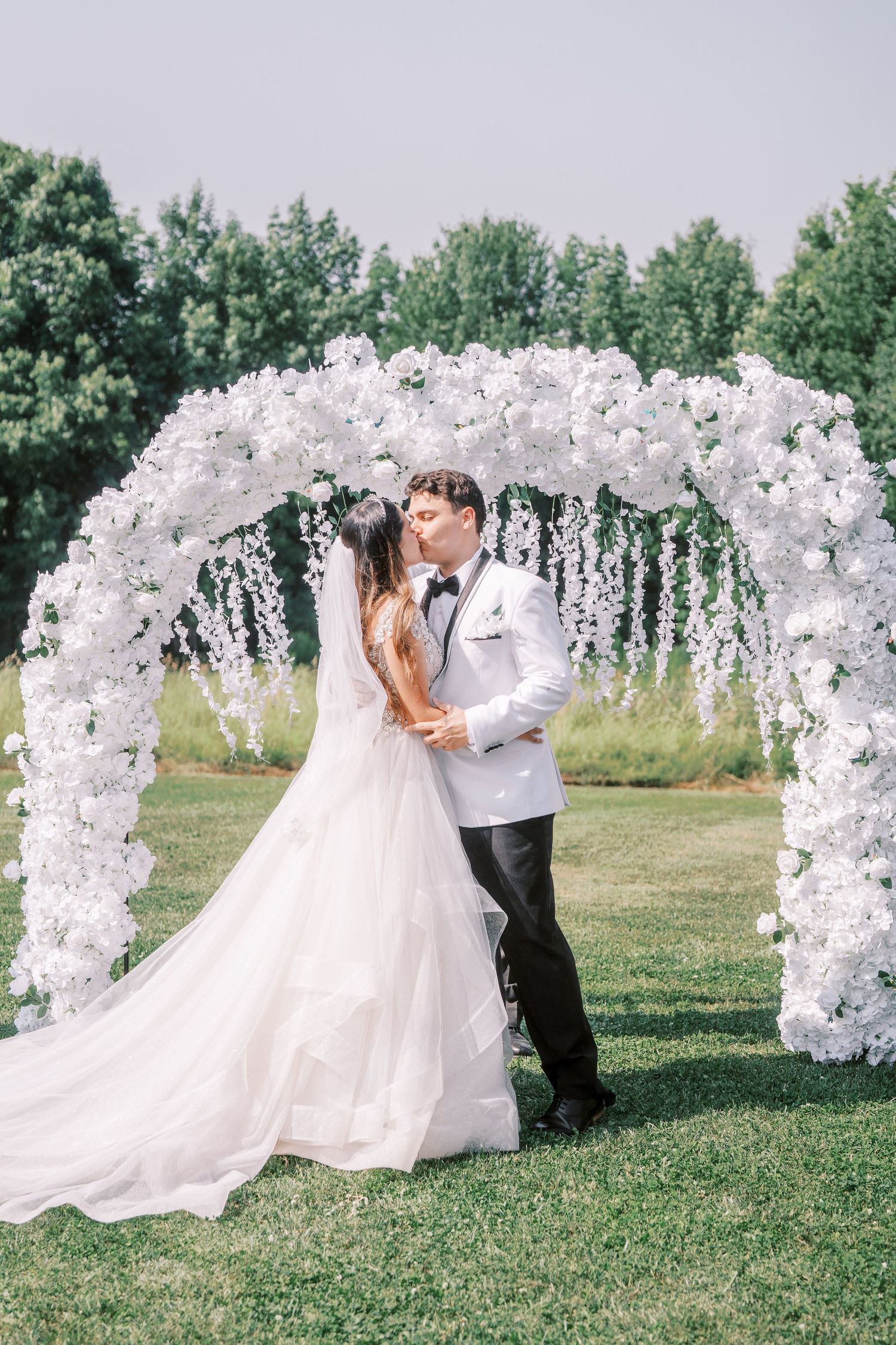 A wedding ceremony setup with a gorgeous white floral arch on a grassy lawn framed by trees in the background.