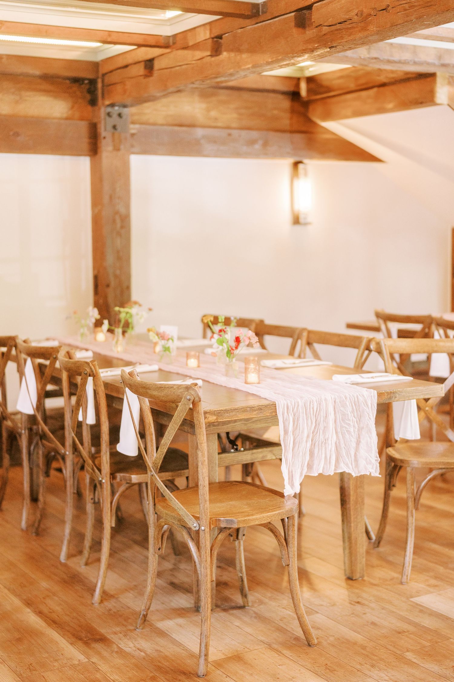 Rustic wedding reception venue features wooden cross-back chairs and white linens under exposed beam ceiling.