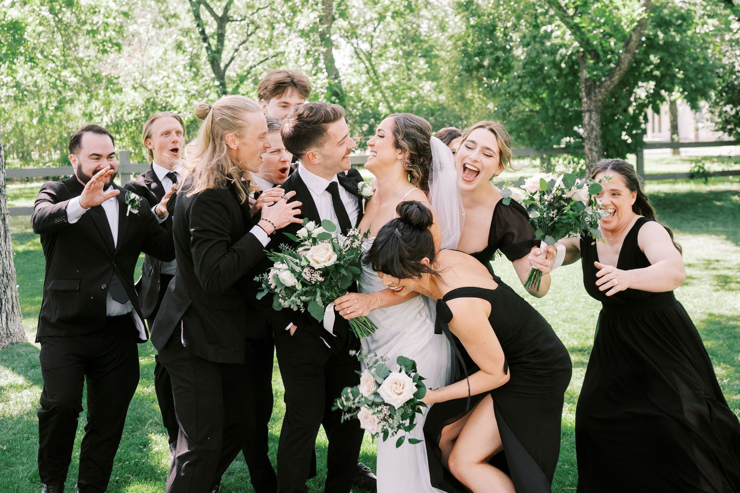 A joyful wedding party in black attire laughs together while posing outdoors in a sunlit garden setting.