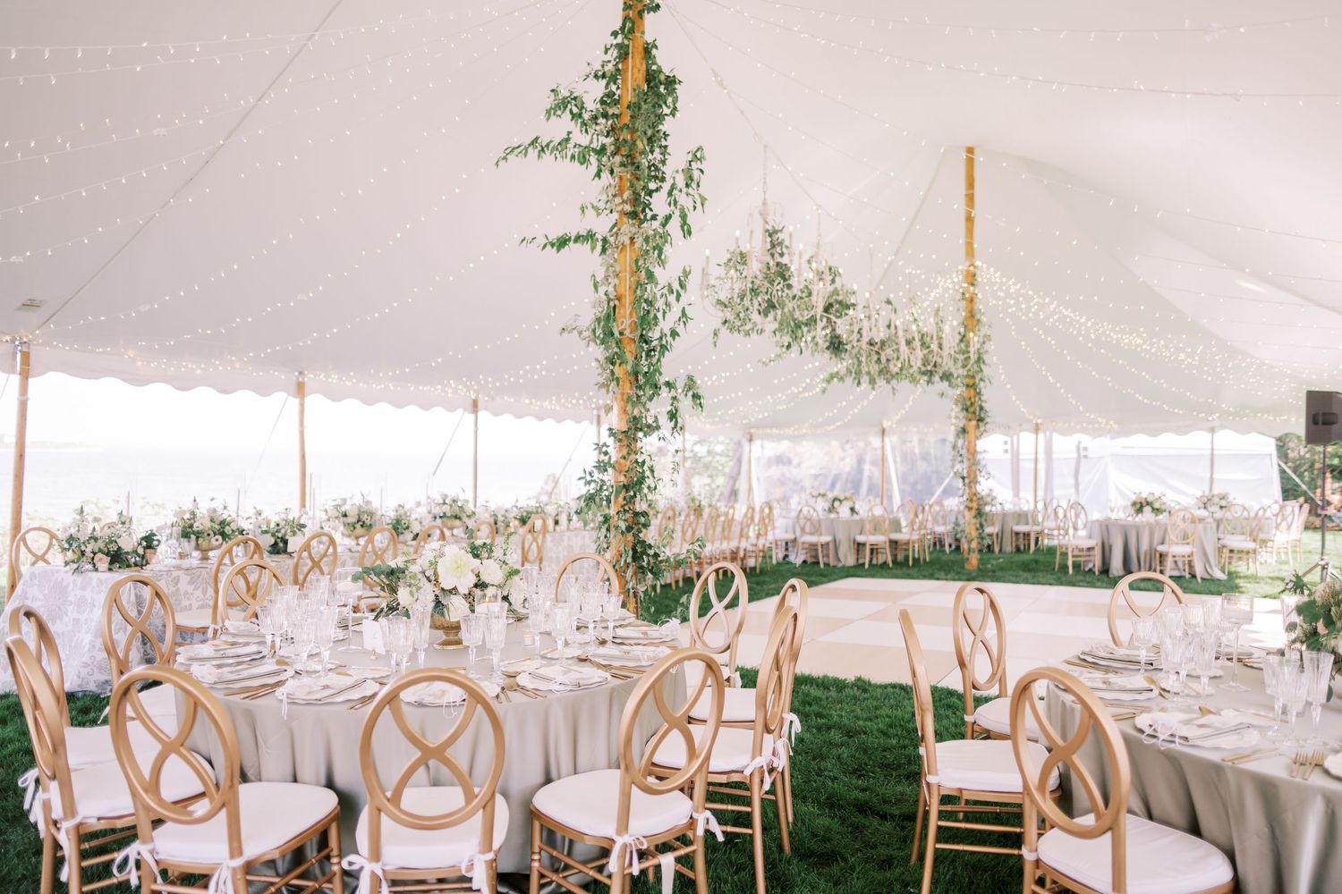 An elegant outdoor wedding reception tent with white linens, wooden crossback chairs and greenery-wrapped support poles.