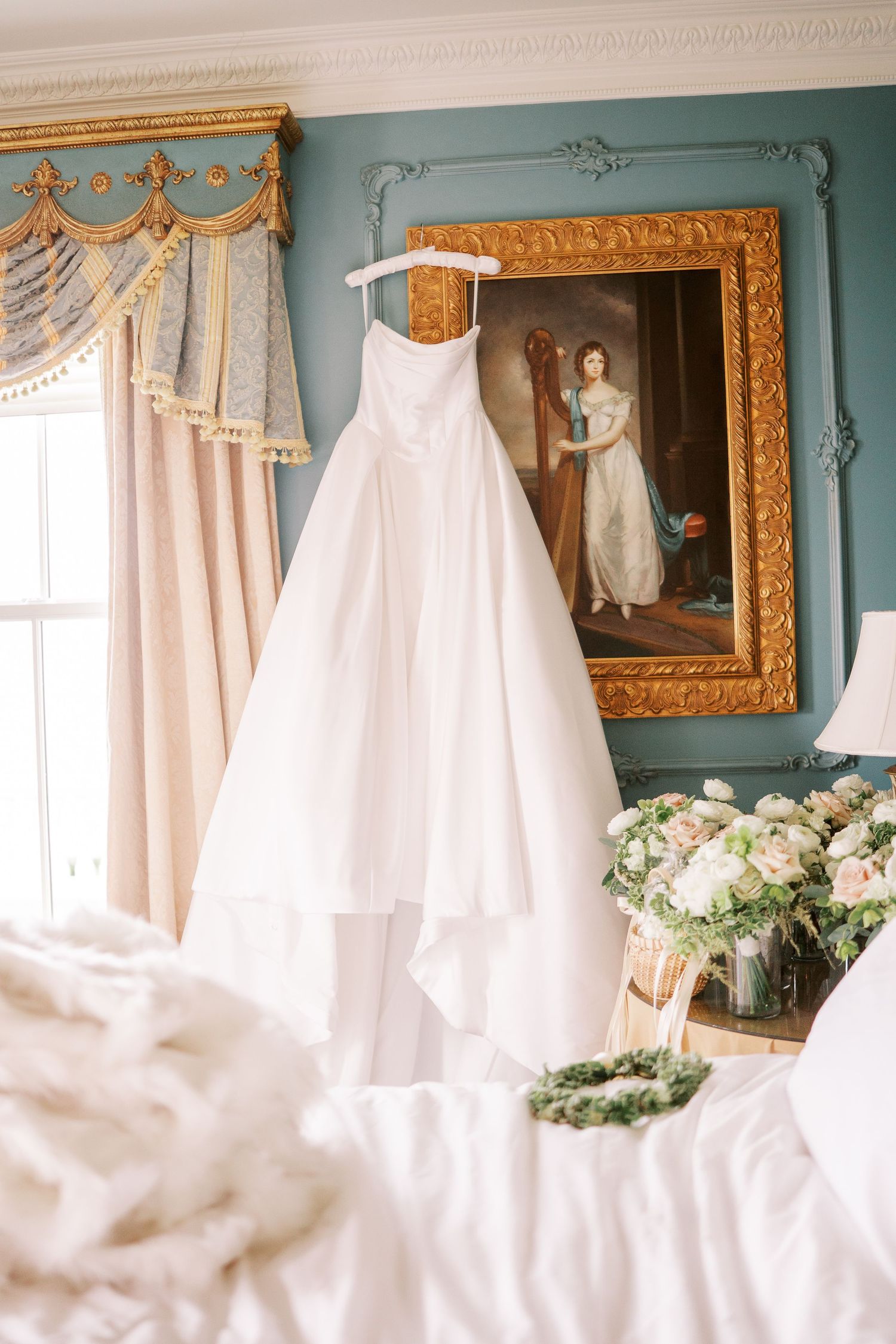 A white wedding dress hangs in an elegant blue room with ornate gold framed artwork and floral arrangements.