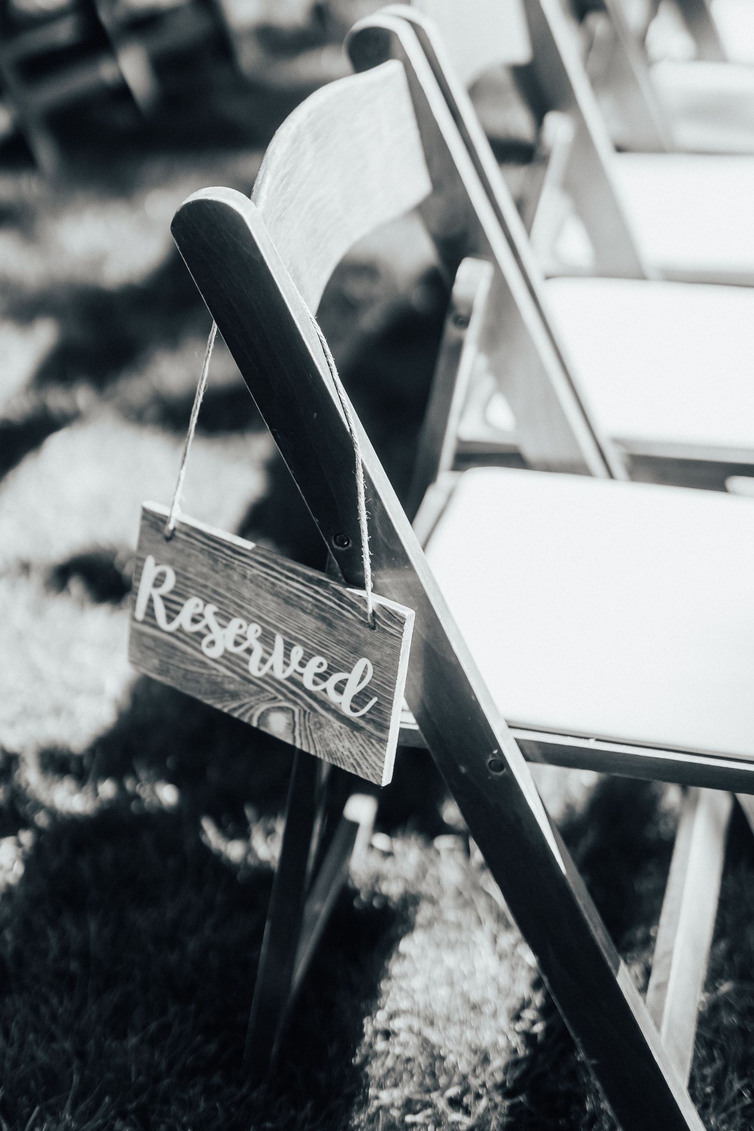 Black and white photo of a wooden folding chair with a rustic 'Reserved' sign hanging on its back at an outdoor event.