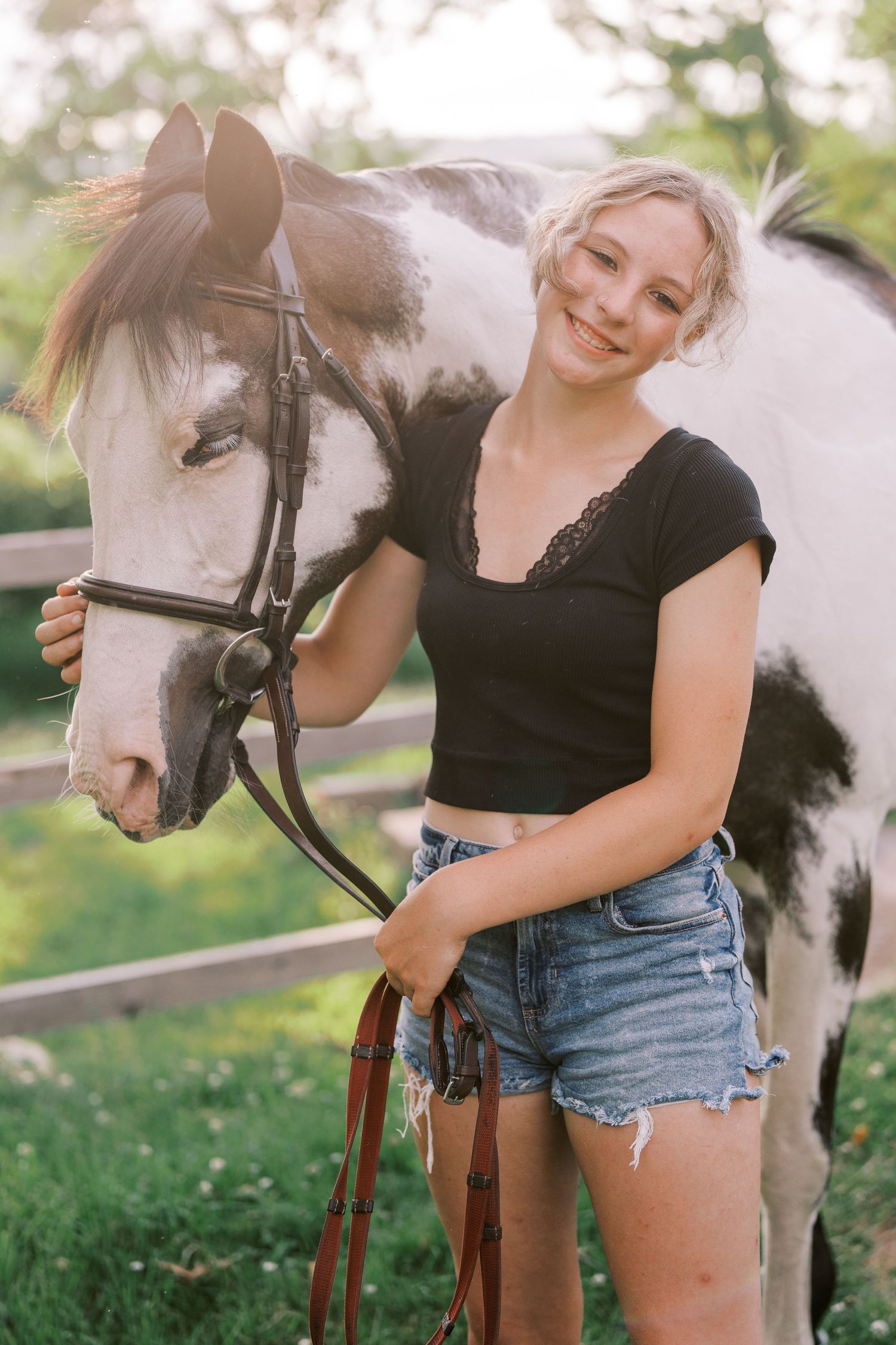 A paint horse with white and dark markings wearing a bridle stands with someone in casual summer attire.