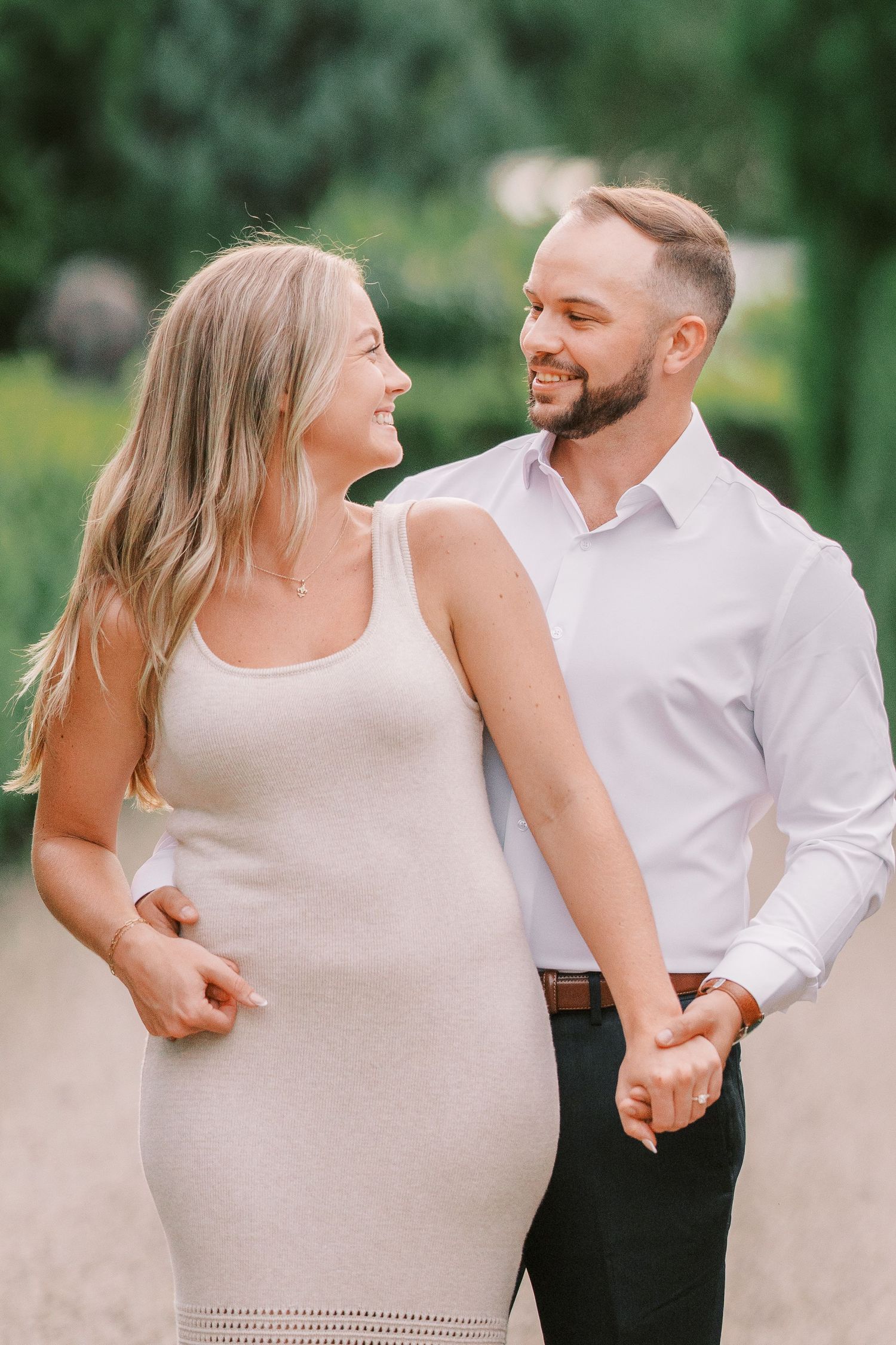 A happy couple walking on a path outdoors, dressed in cream and white, laughing and holding hands in a romantic moment.