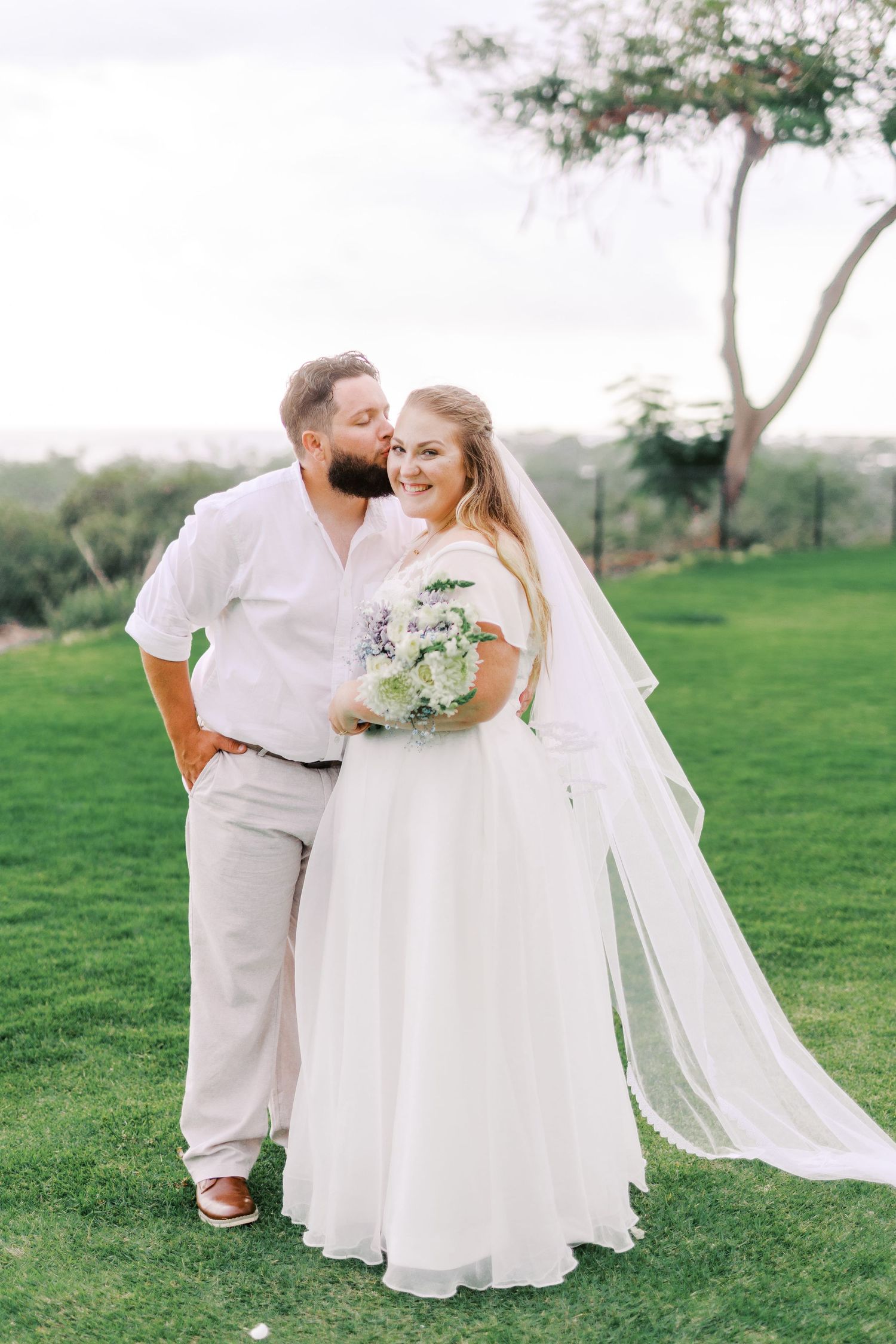 A joyful couple poses on a grassy hill during their outdoor wedding photoshoot.