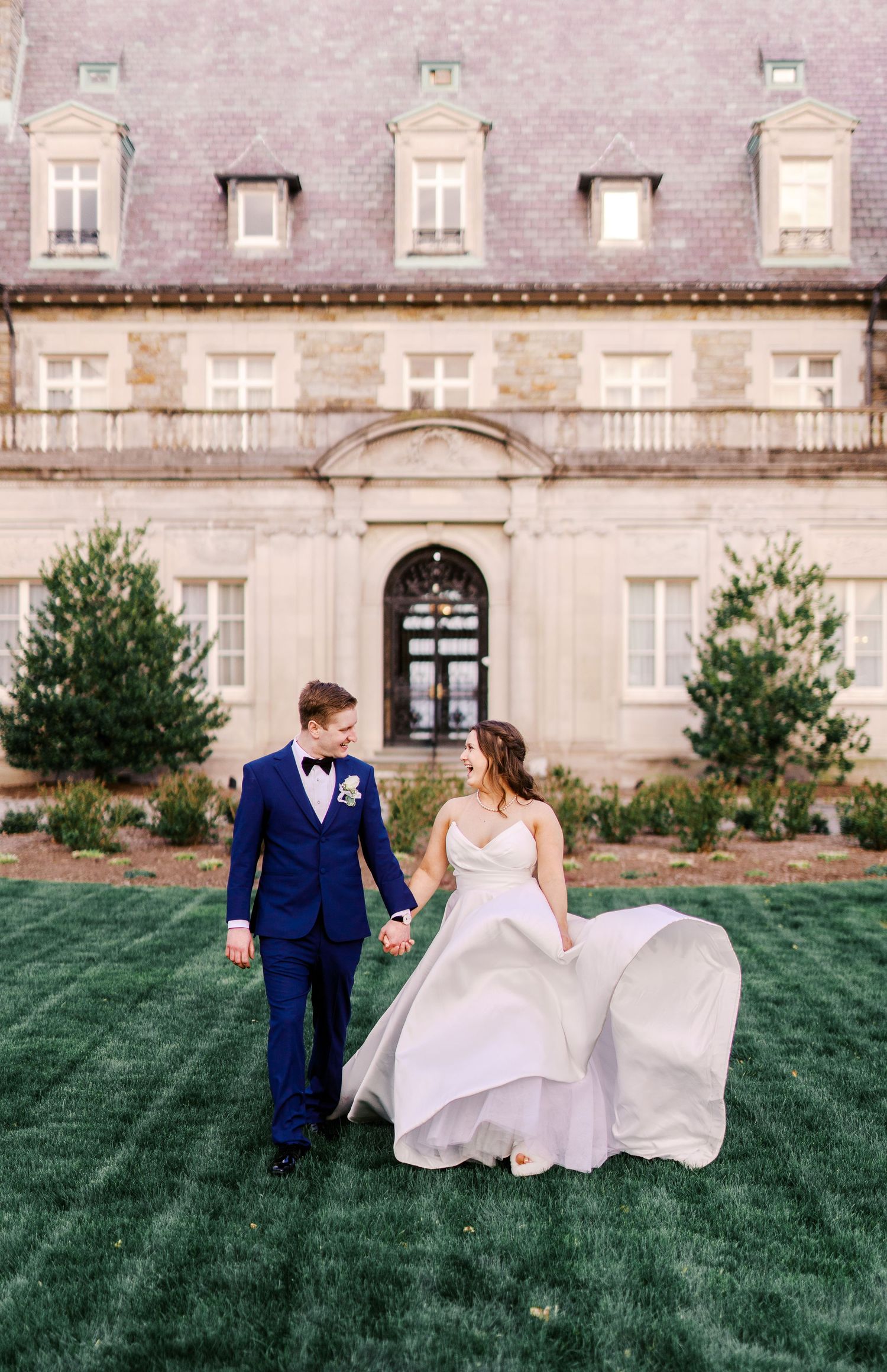 A couple in wedding attire holding hands on a lawn in front of an elegant stone mansion with dormer windows.