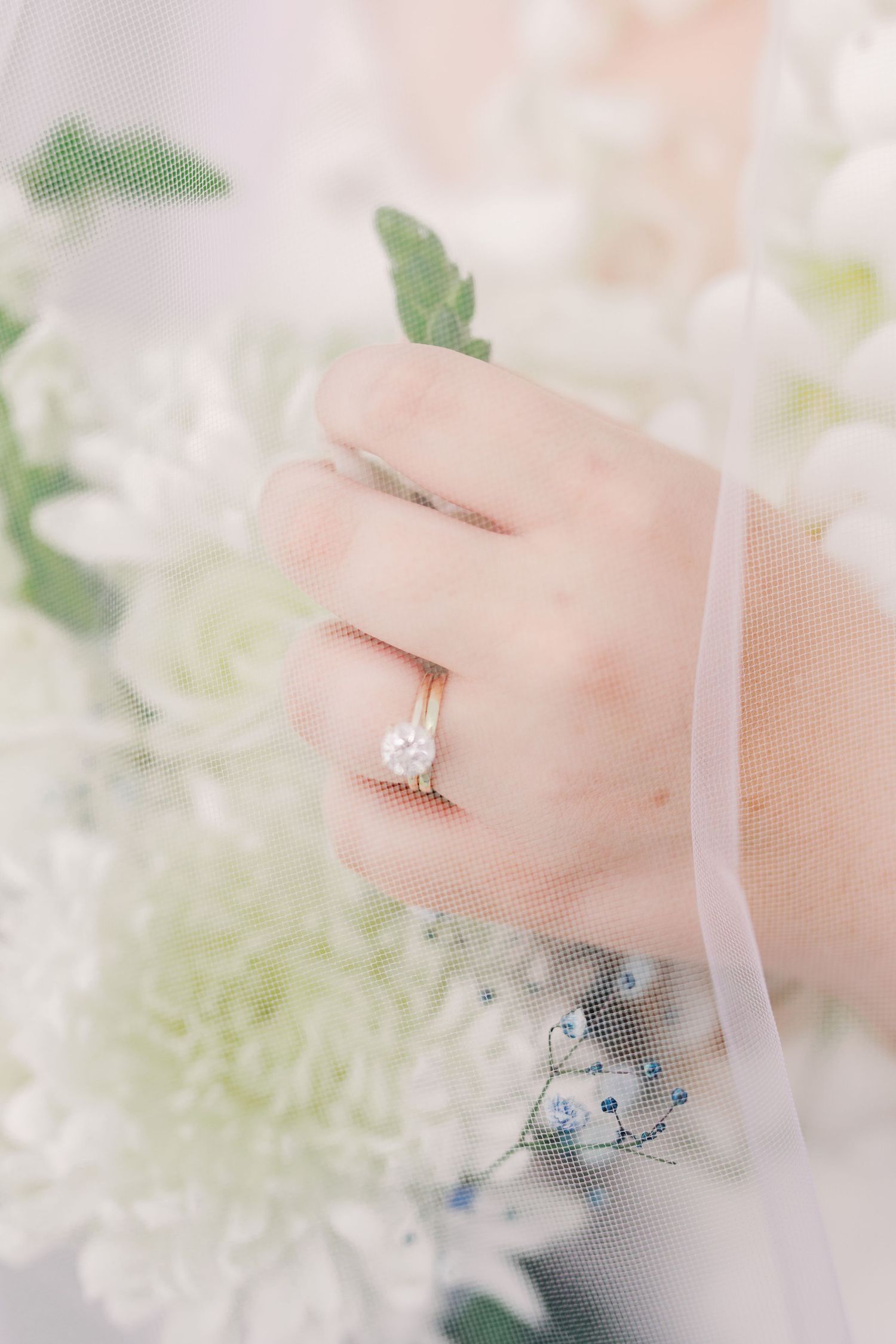 A delicate engagement ring sparkles against soft white flowers and tulle veil.