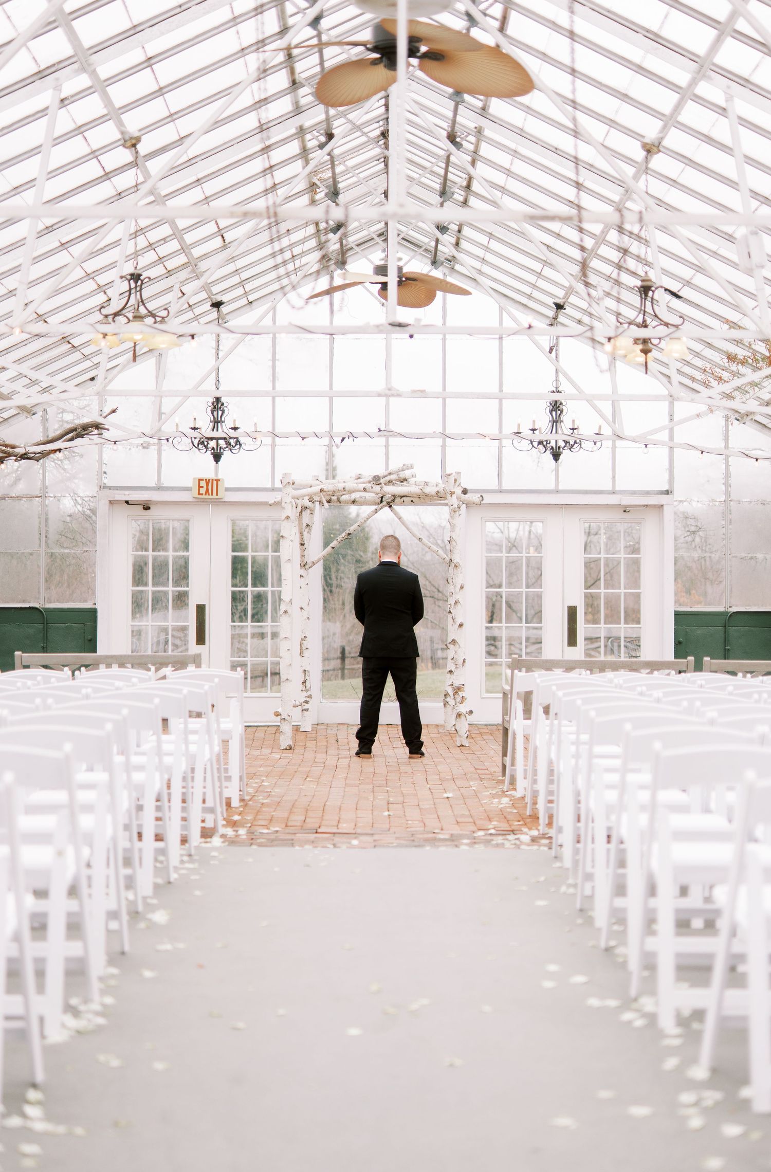 A minimalist greenhouse wedding venue with white chairs and crystal chandeliers awaits a ceremony in dramatic lighting.