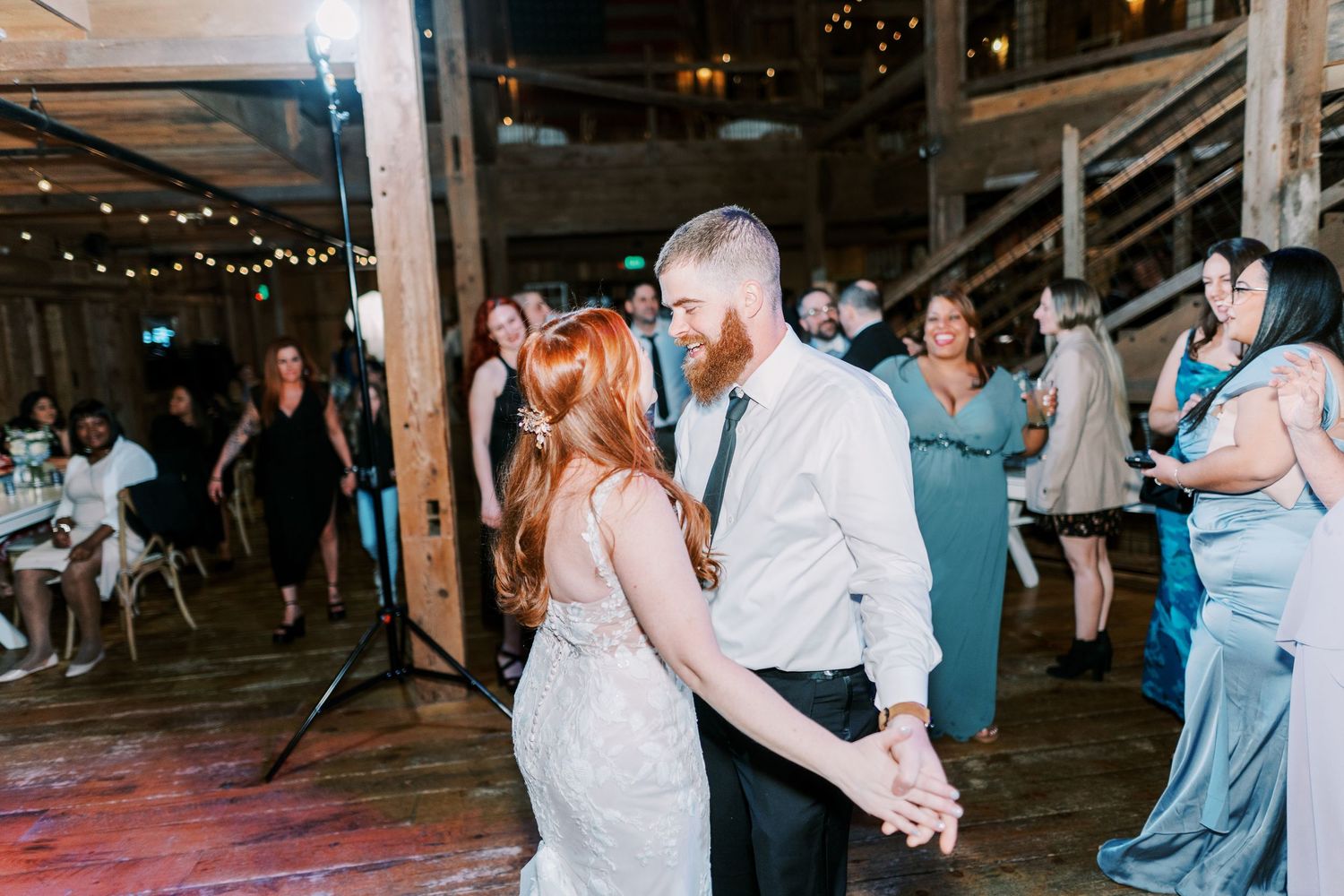 Couple dancing together at an indoor wedding reception with string lights and guests in the background.