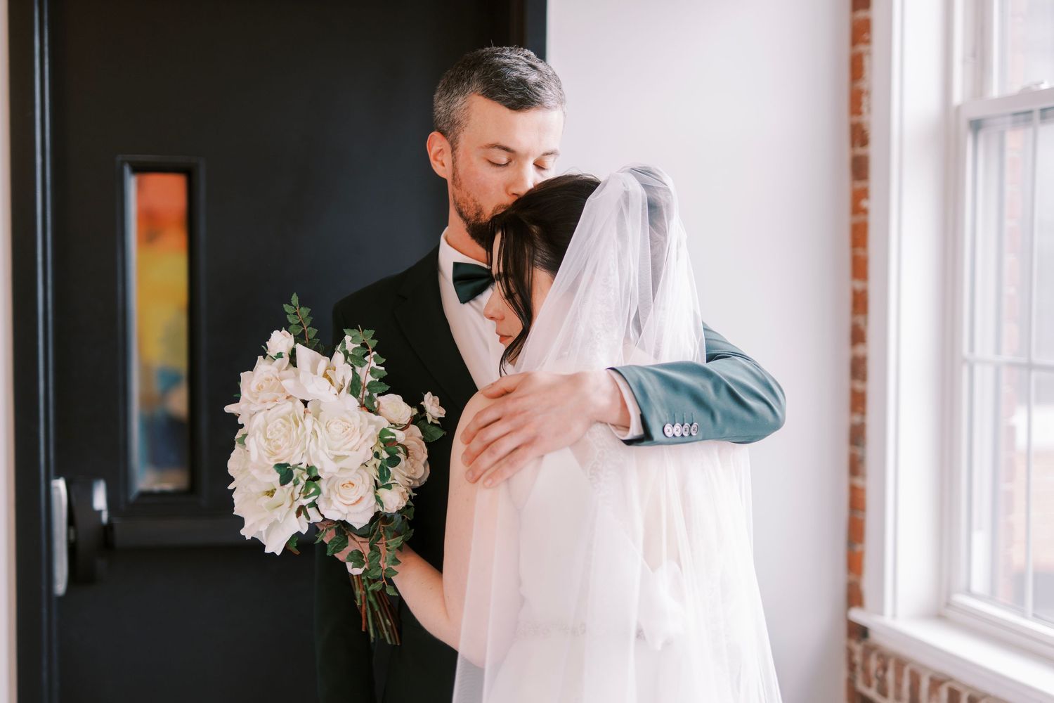 A tender moment between newlyweds embracing near a window with a white floral bouquet.