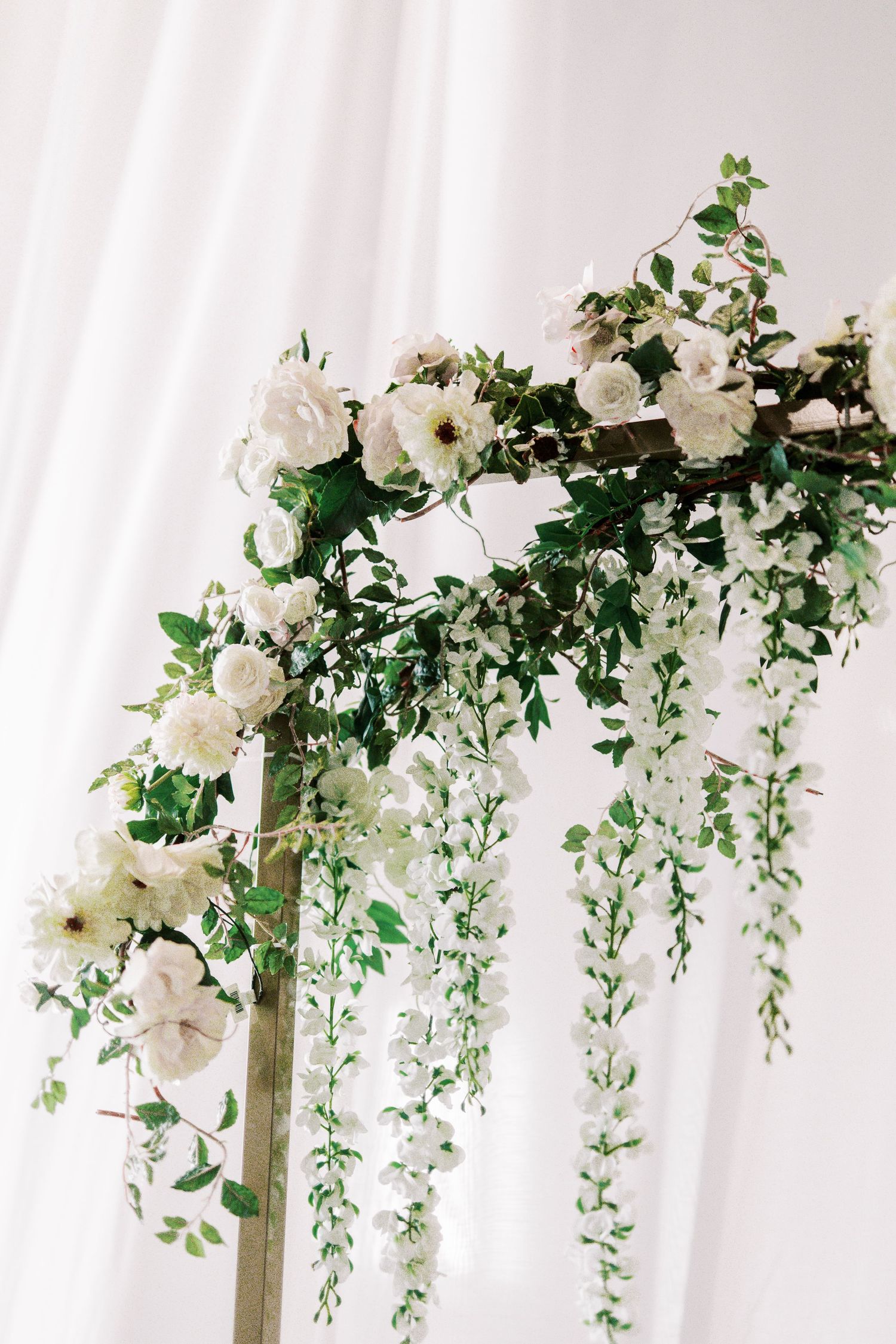 Close-up of white roses and cascading greenery on a wedding arch against white backdrop.