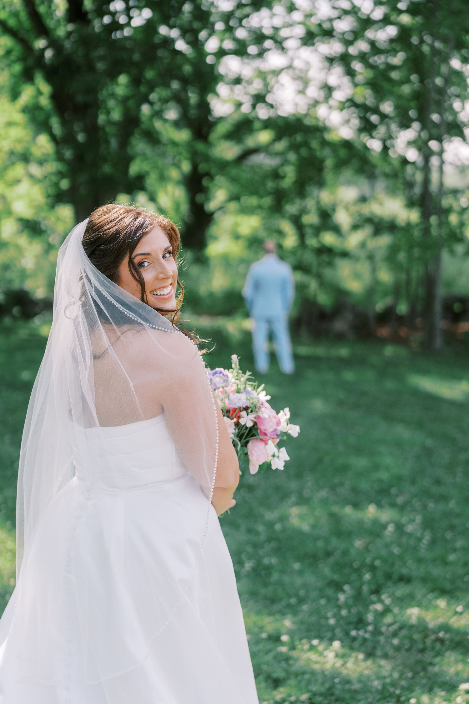 A beautiful bride in a white wedding dress and veil stands in a lush green garden holding a pink and white bouquet.
