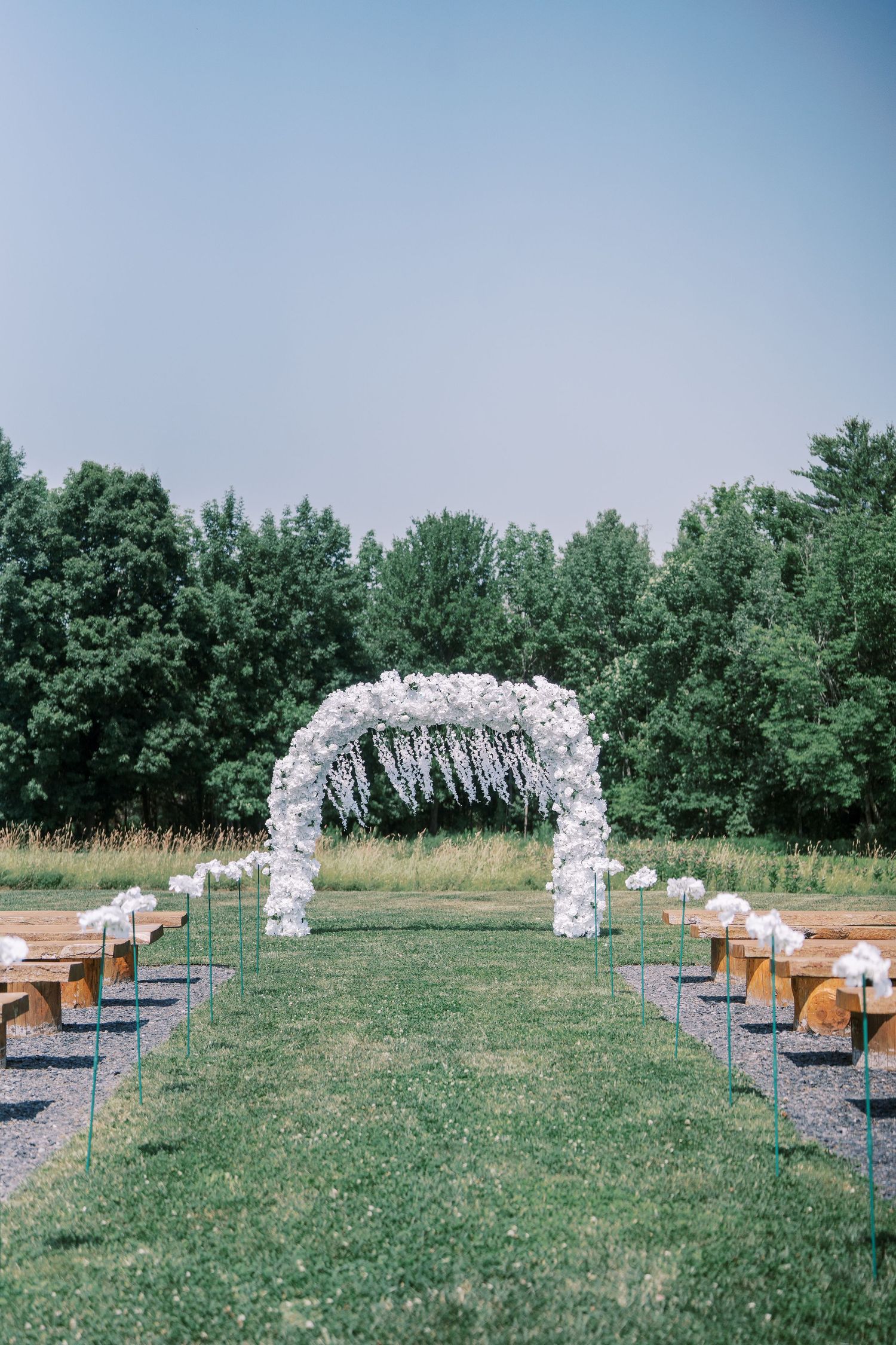Outdoor wedding ceremony setup with wooden benches and white floral arches on grassy field surrounded by trees.