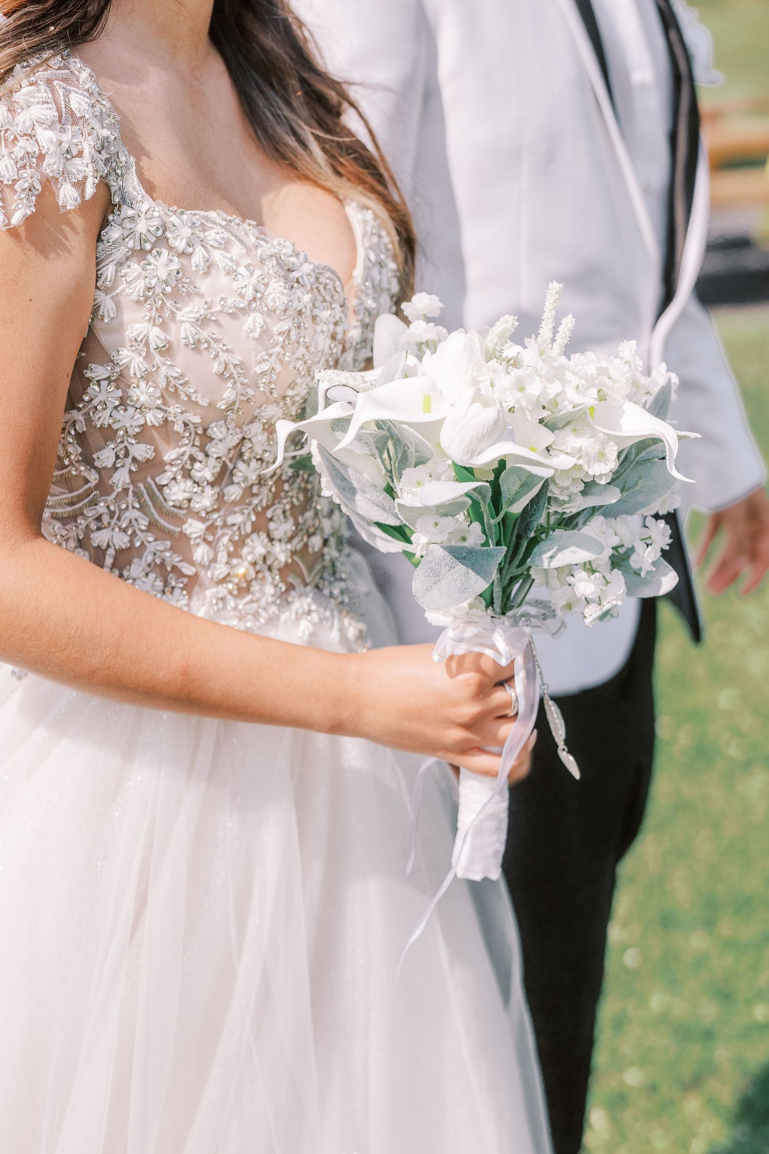 Three people in formal wedding attire holding delicate white floral bouquets with greenery during an outdoor ceremony.