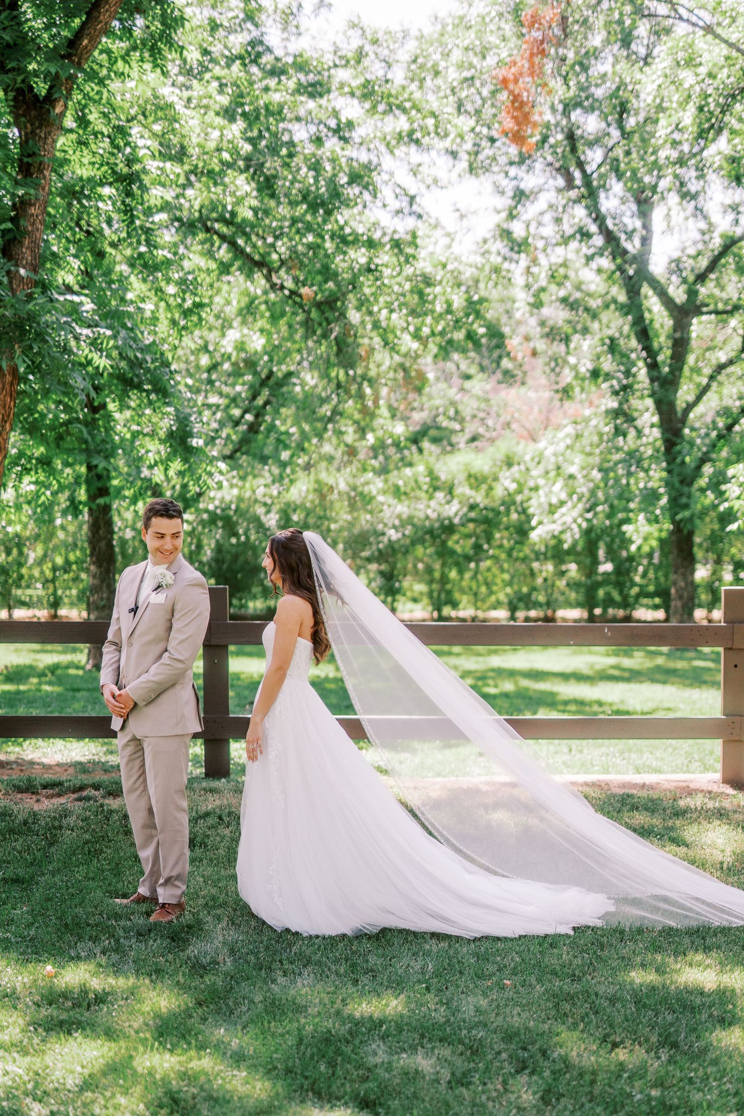 Couple in wedding attire standing outdoors in a lush green garden setting with a long flowing veil in the sunlight.
