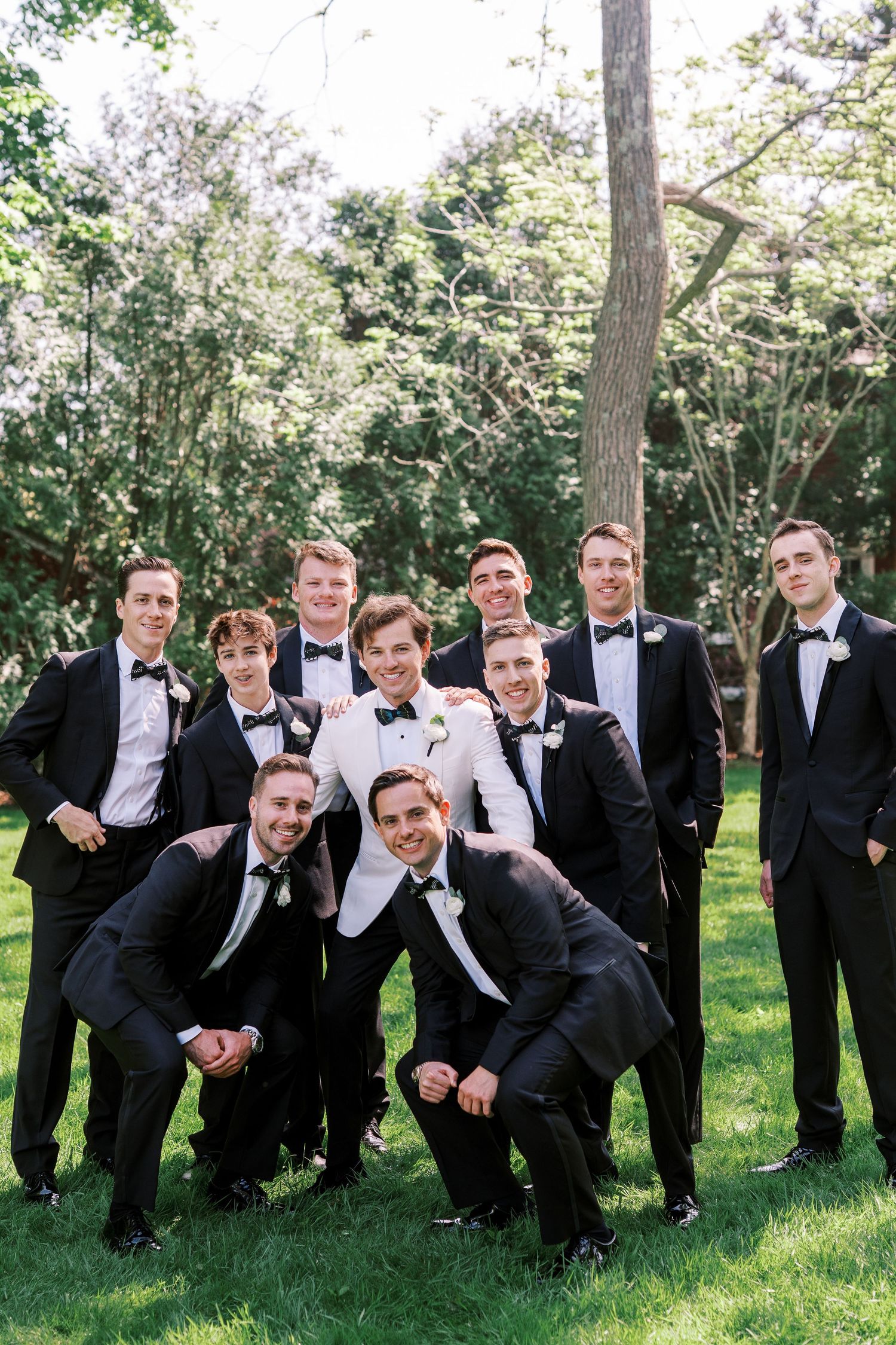A group in formal black tuxedos poses playfully together on grass in a creative line formation near trees.