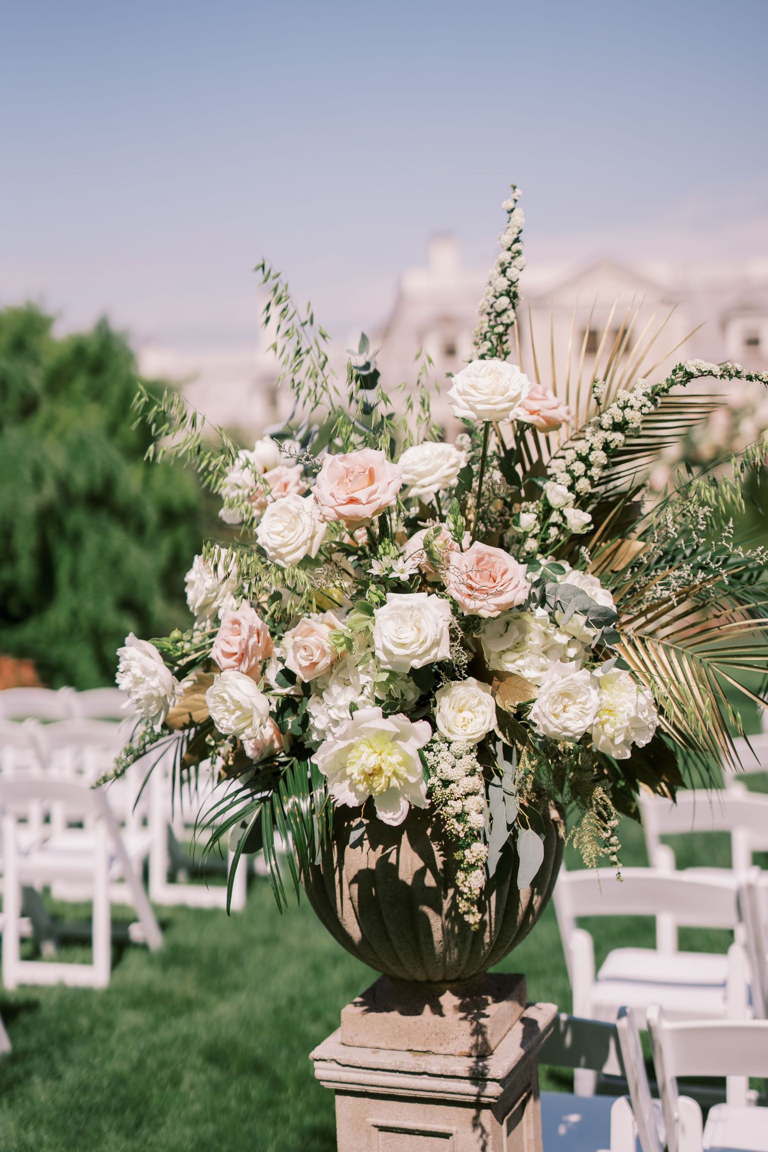 Elegant floral arrangement with pink and white roses in dark urn on pedestal with white chairs and green lawn background.