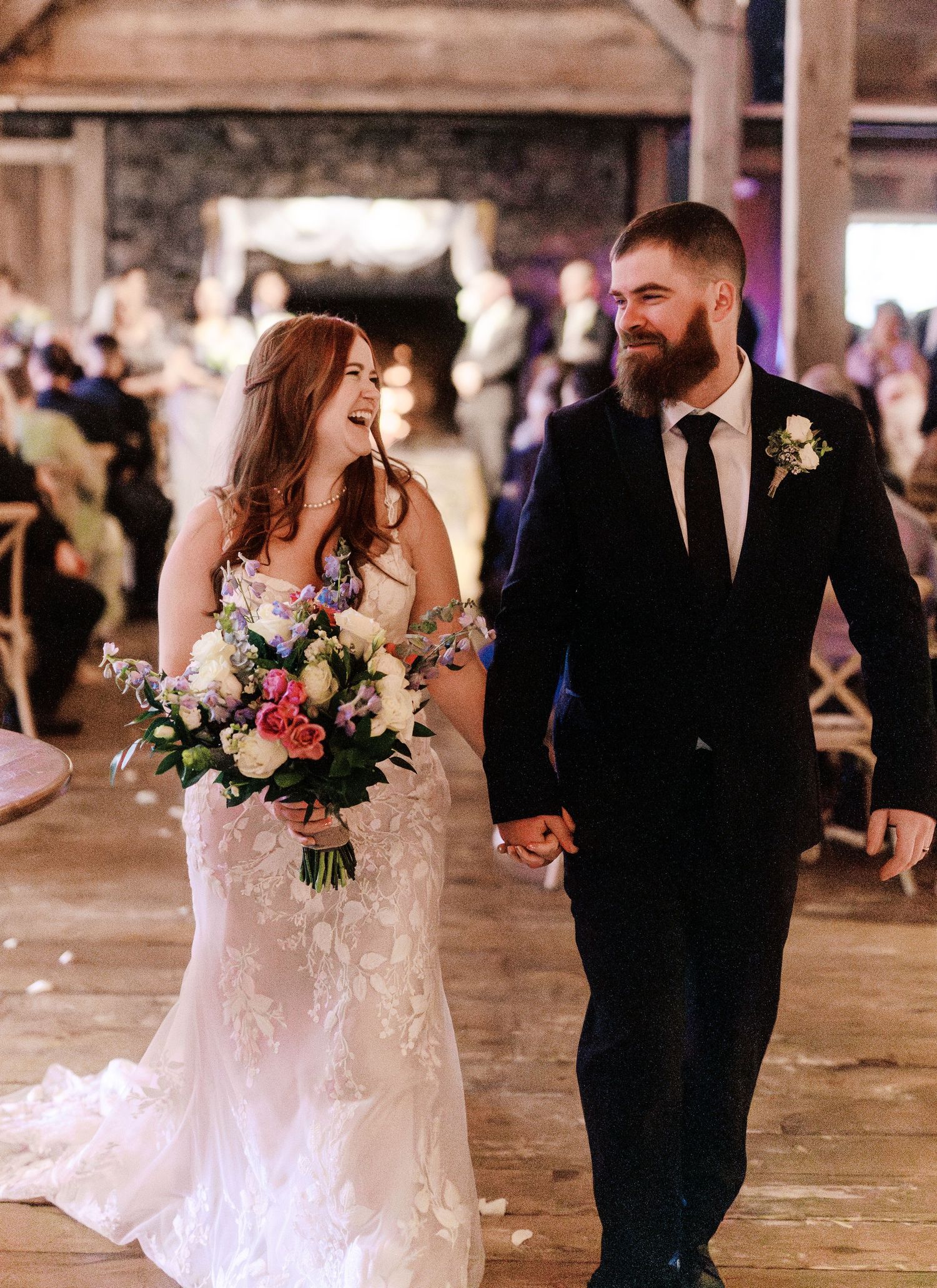 A joyful couple walking hand in hand at their rustic barn wedding celebration, holding a pink and white floral bouquet.