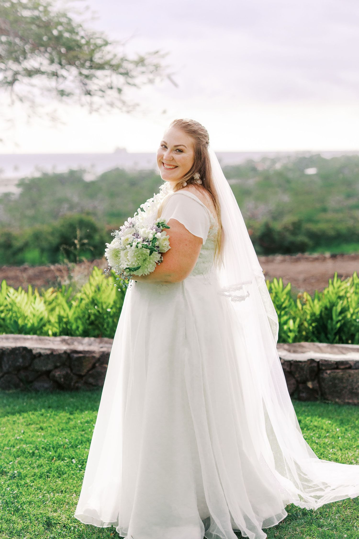 Bride holding white bouquet poses on lawn with scenic valley view.
