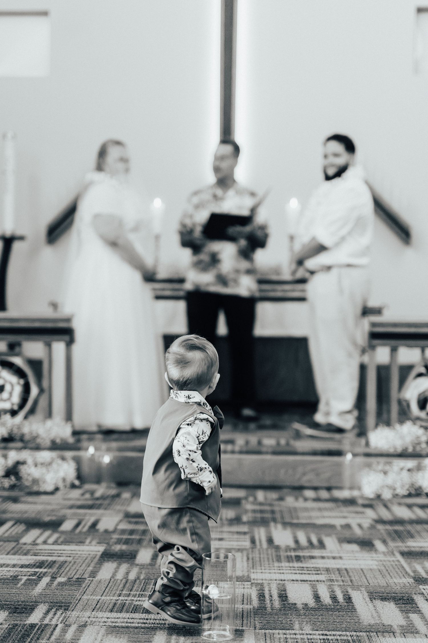 A black and white photo of a small child watching a wedding ceremony from the aisle.