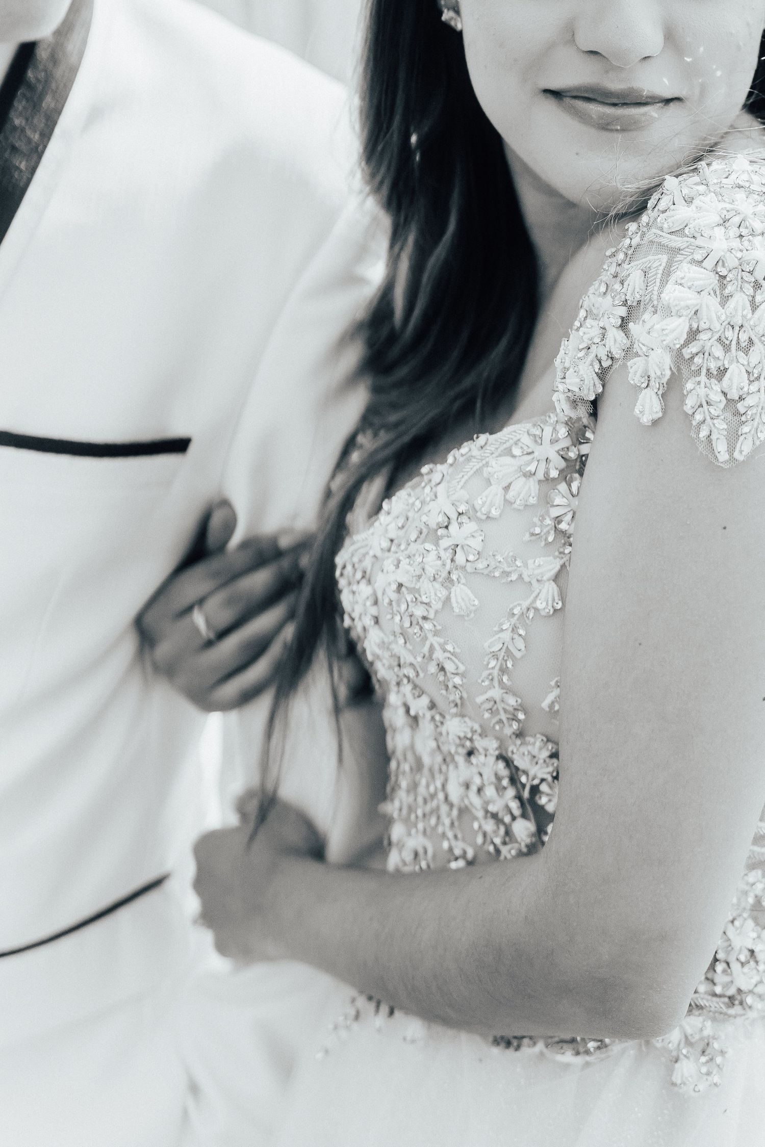 Black and white wedding photo showing intricate lace detail on a wedding dress and part of a white tuxedo jacket.