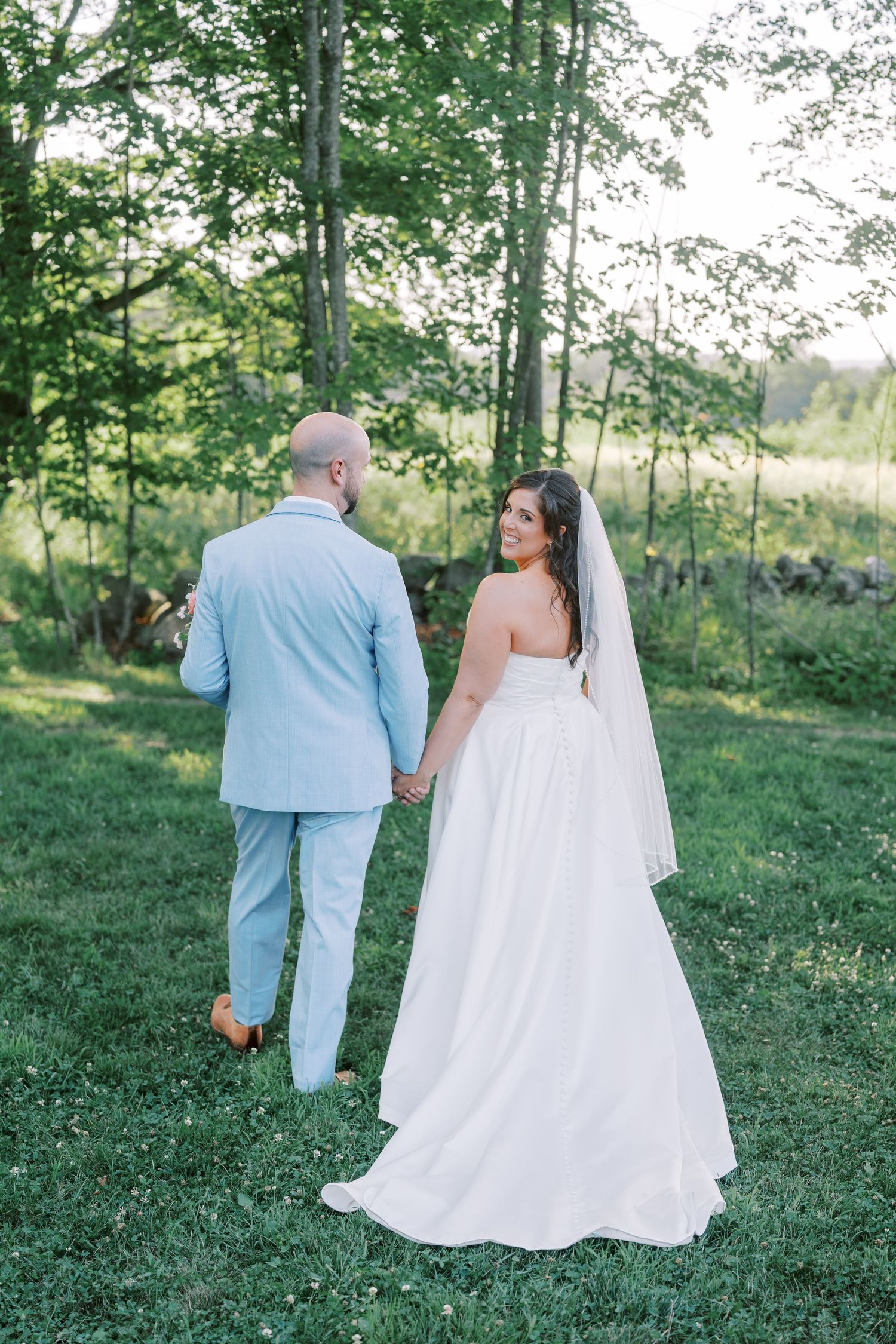 Couple walking hand in hand through a sunlit grassy field in wedding attire.