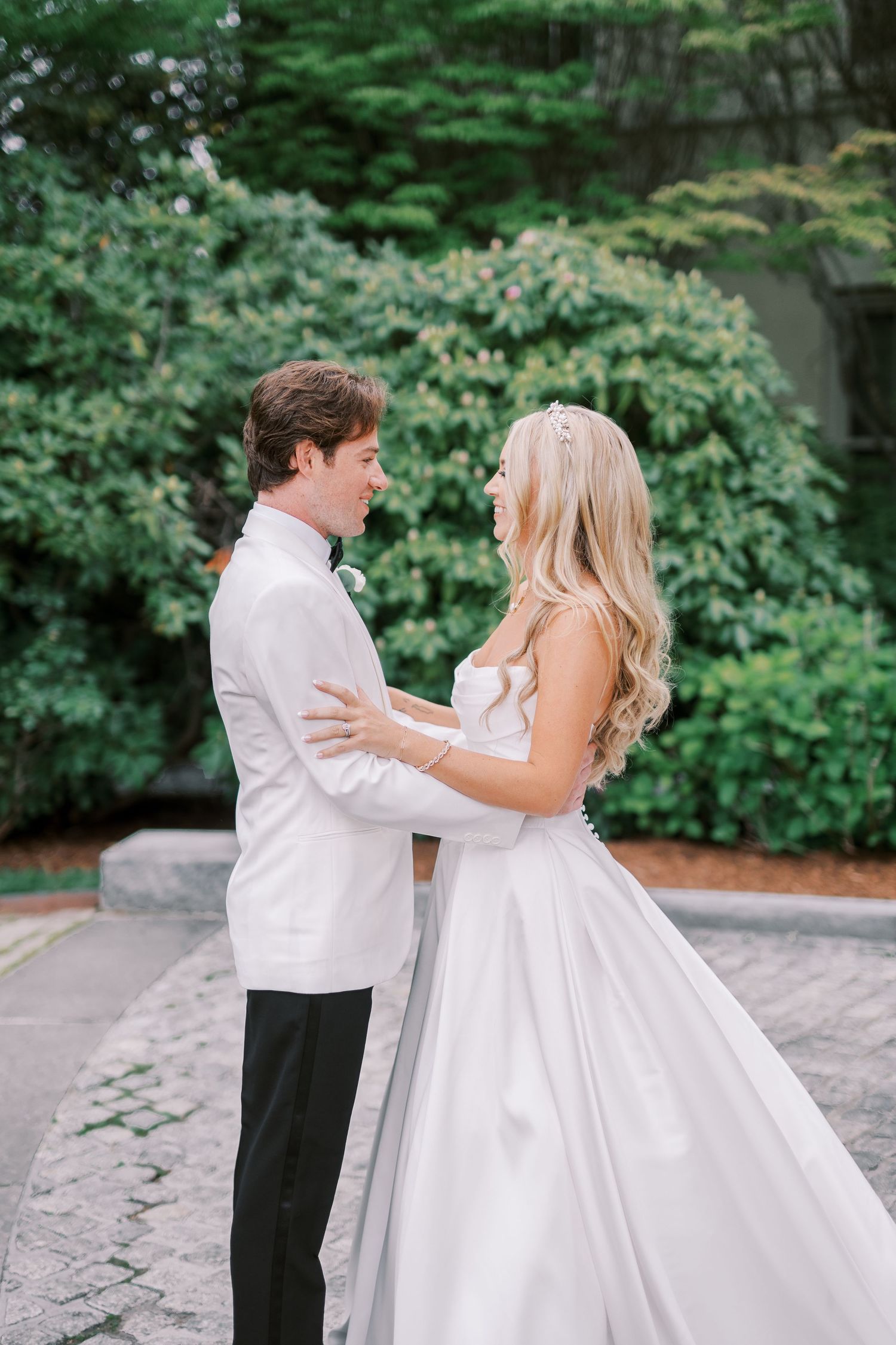A romantic wedding couple in white attire dance together on a stone patio surrounded by lush green foliage.